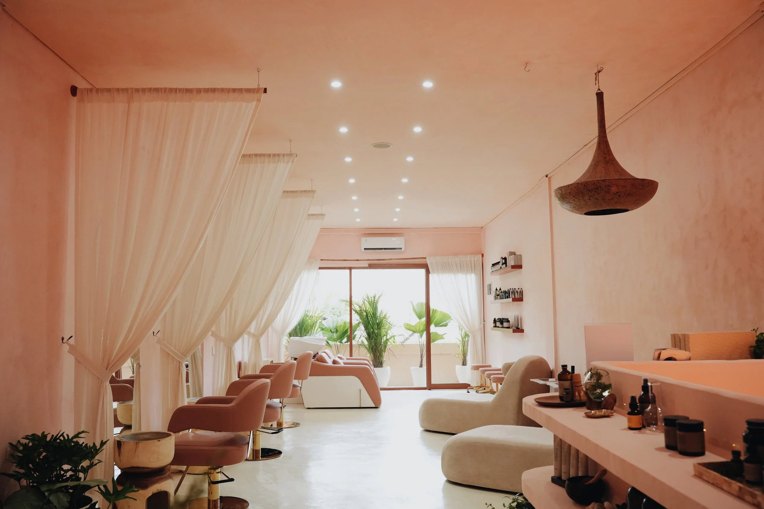 Interior of a cozy, well-lit spa or lounge area with light-colored walls and ceiling, beige chairs with pink cushions, white curtains, a chandelier, and large windows with outdoor plants visible.