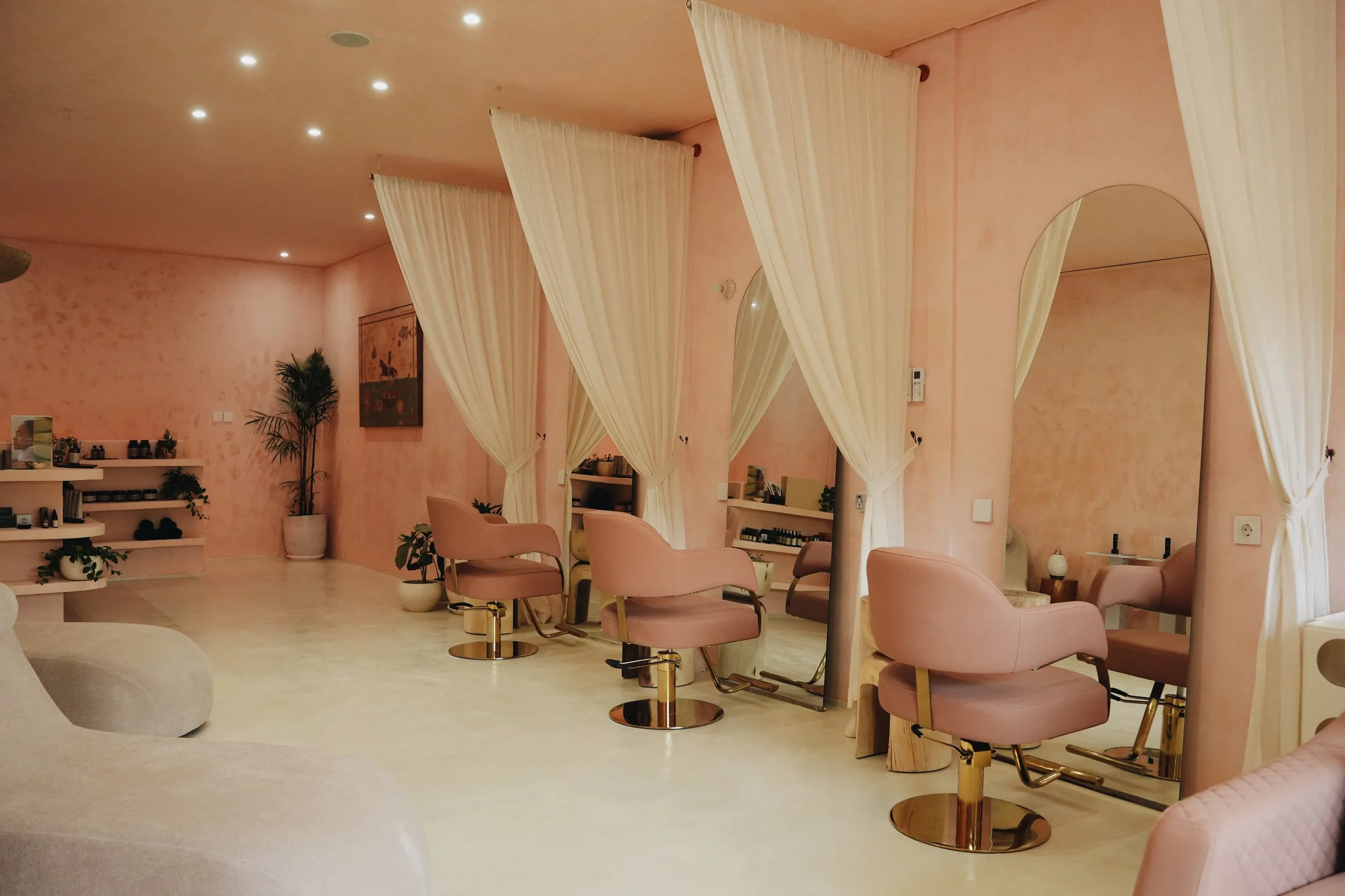 A salon interior with pink walls and matching pink chairs, white curtains, mirrors, potted plants, and a shelf with hair care products.