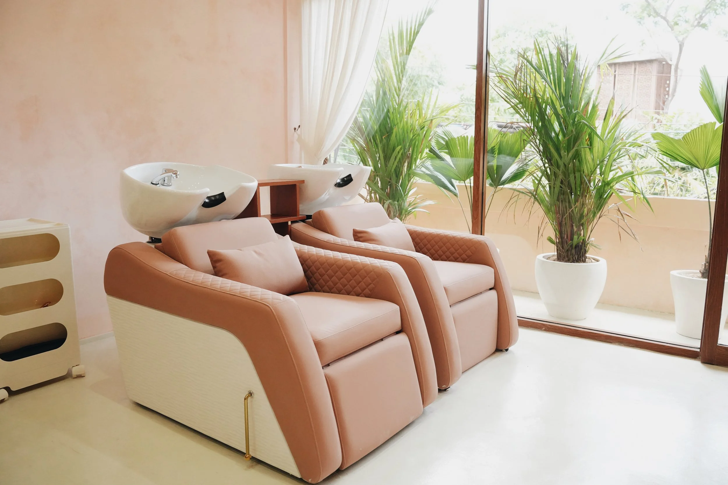 A hair salon or spa room with two white shampoo bowls, a brown couch with pillows, large potted plants near a glass window, and a beige wall with a white curtain.