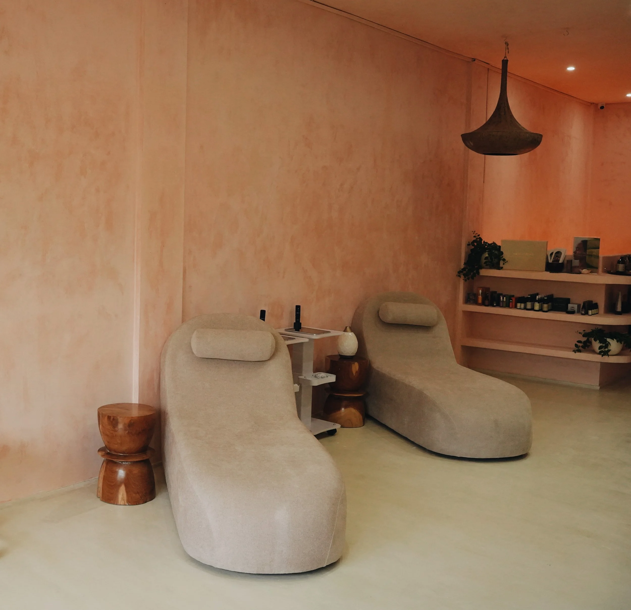 Interior with peach-colored walls, a hanging lamp, beige upholstered furniture, a small wooden side table, and a white shelf with books and photos.