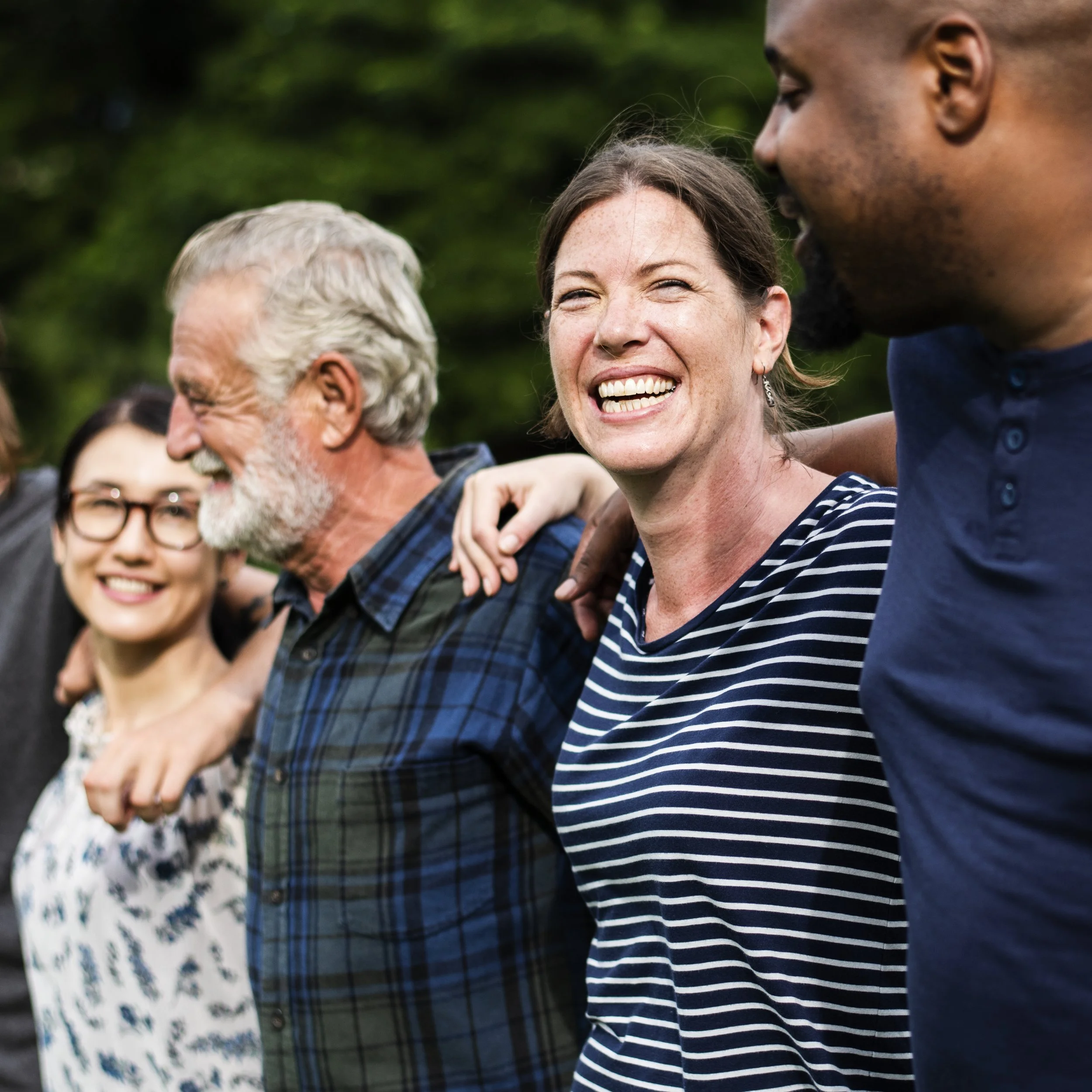 A group of diverse people, including an older man and younger adults, standing outdoors with arms around each other's shoulders, smiling and enjoying the moment.