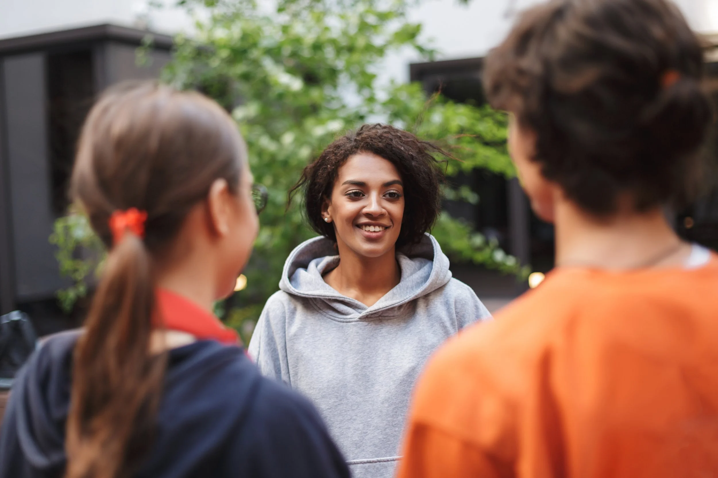 A woman smiling and talking outdoors with two children, one girl with brown hair tied back, and one boy with short hair, against a background of greenery and dark outdoor structures.