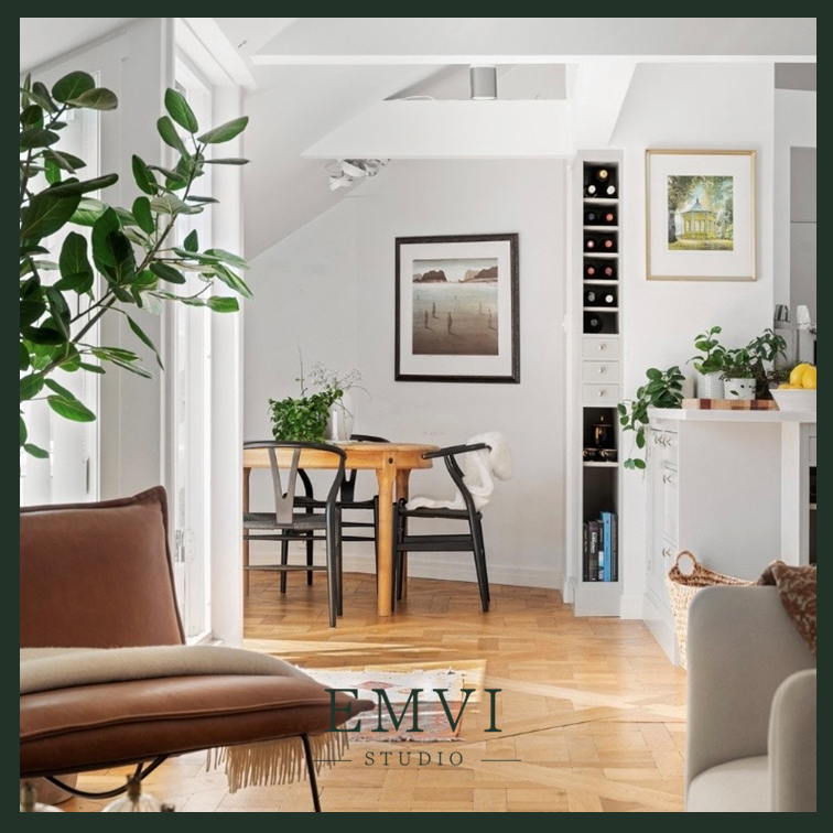 Bright, cozy living room with white walls and wood flooring. Features a brown sofa, a round dining table with four chairs, and framed artwork on the walls. A tall window with white curtains lets in natural light, and there are potted plants and a wine storage unit in the room.