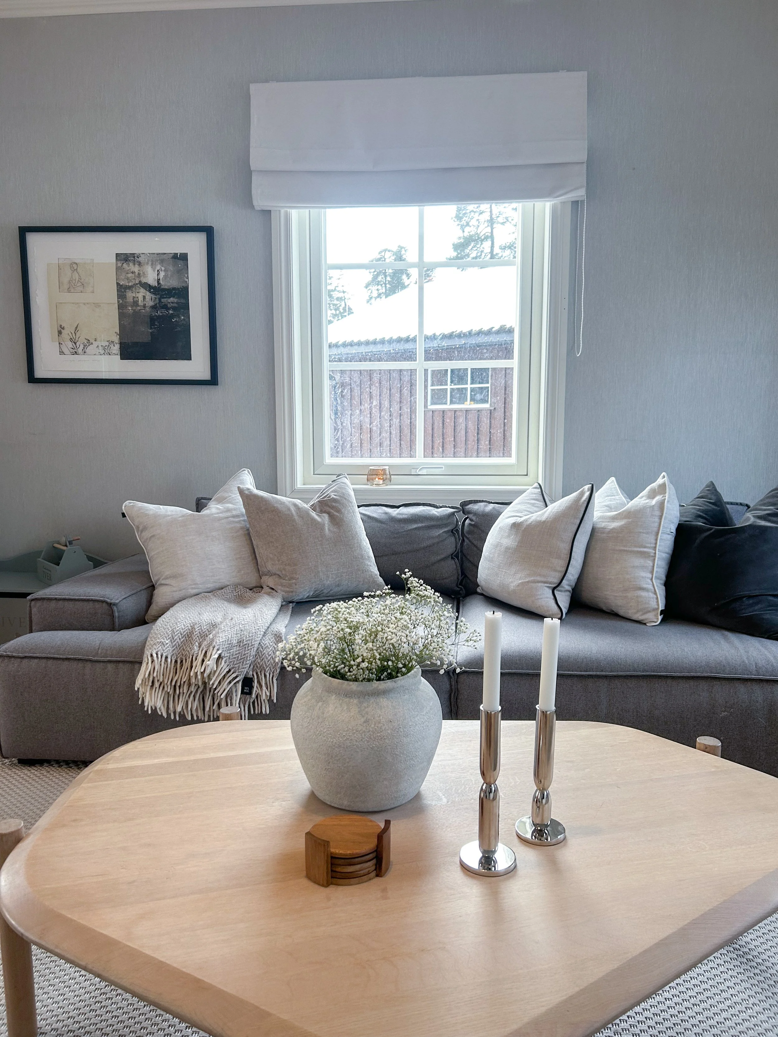 Living room with a gray couch, multiple throw pillows, a light-colored blanket, a wooden coffee table with a white vase of white flowers and two silver candlesticks, and a window with a white blind showing a snowy outdoor scene.