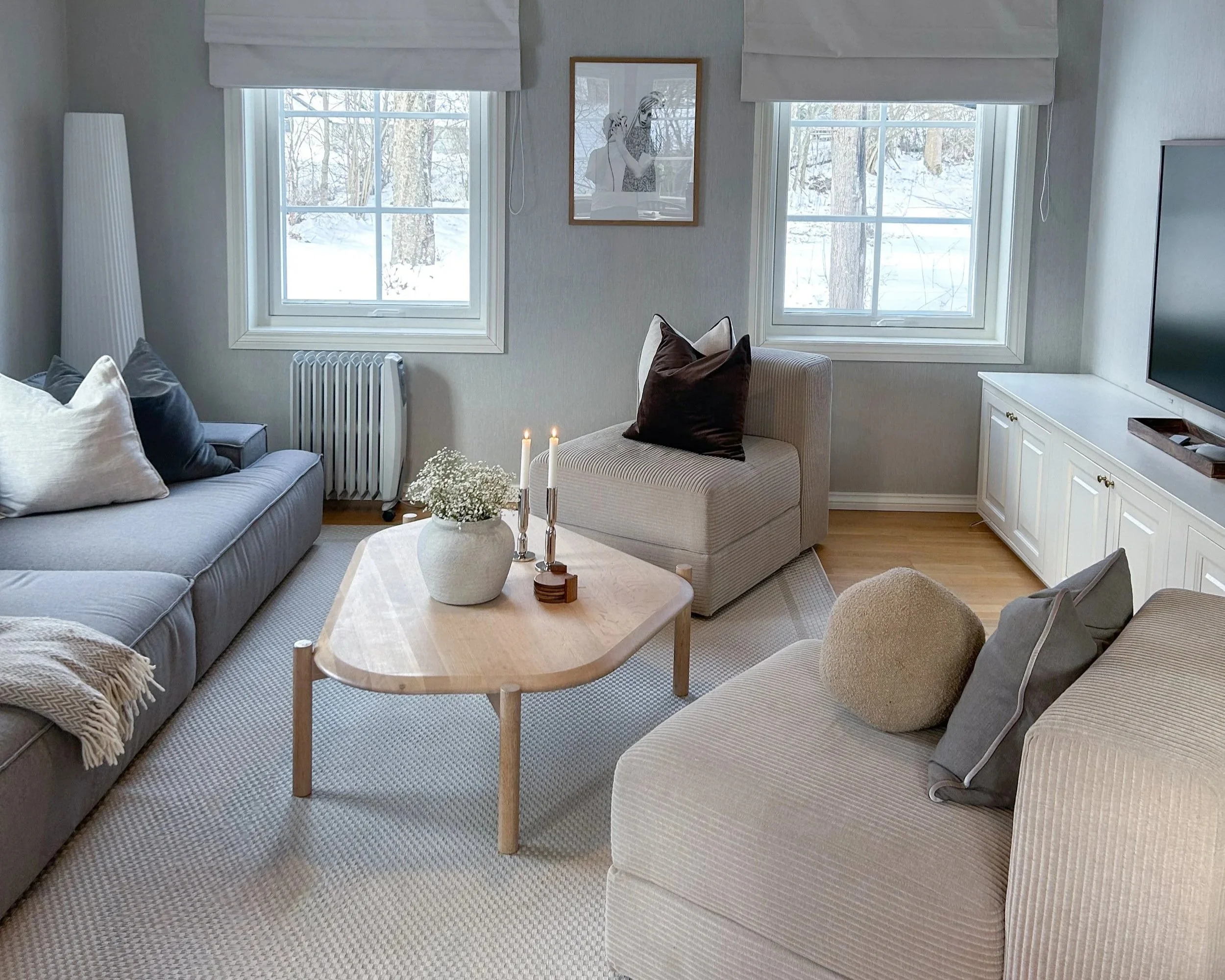 Stue med to vinduer som viser snødekt landskap, sofa, lenestol, TV, tre lysestaker med lys, vase med blomster, og dekorativt bilde på veggen.