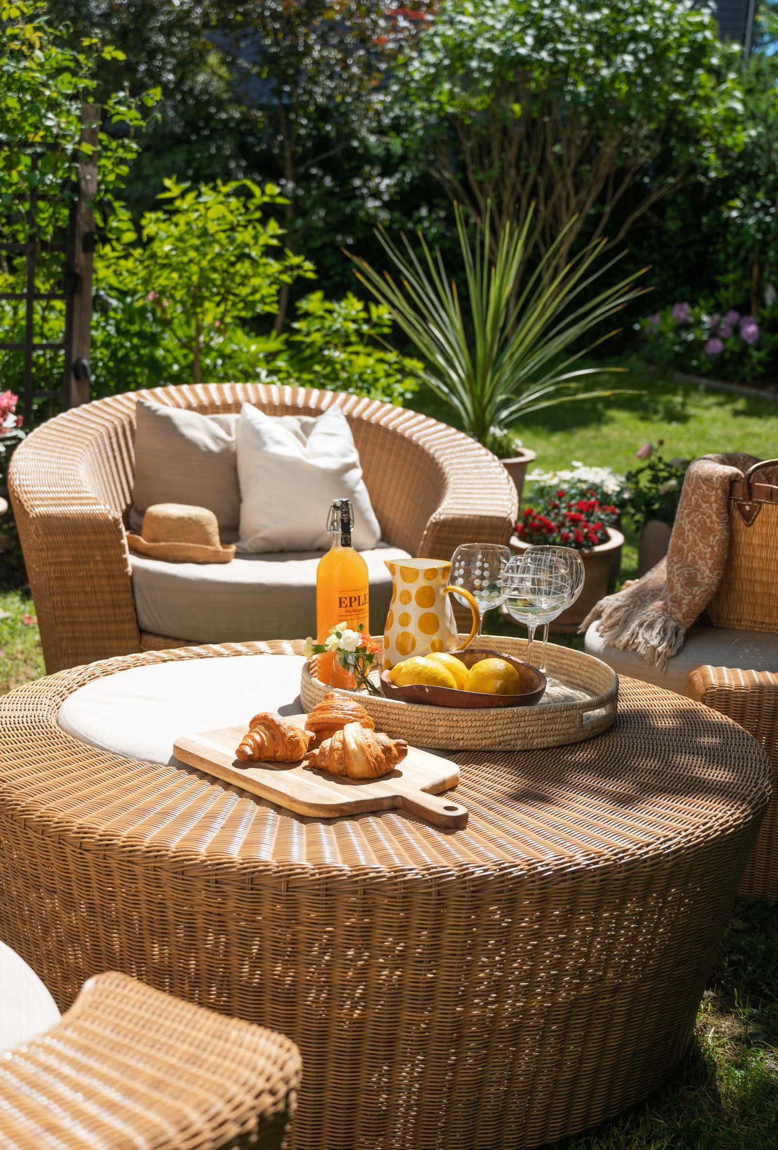 Utendørs hagemøbler med frokost, croissanter, juice, vinflaske, glass og blomster på bord, omringet av grønne planter og blomster
