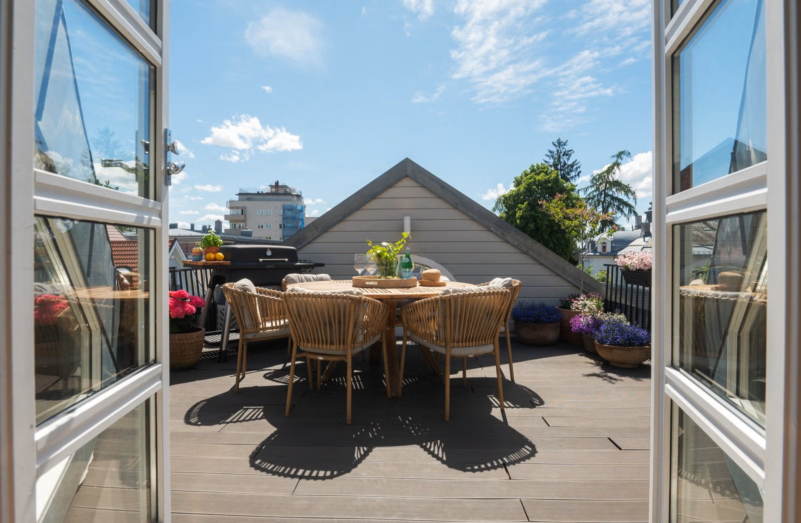 Uteplass med rundt spisebord og seks stoler, omgitt av blomsterpotter med fargerike blomster, sett gjennom utadslående doble glassdører, under blå himmel med noen skyer.