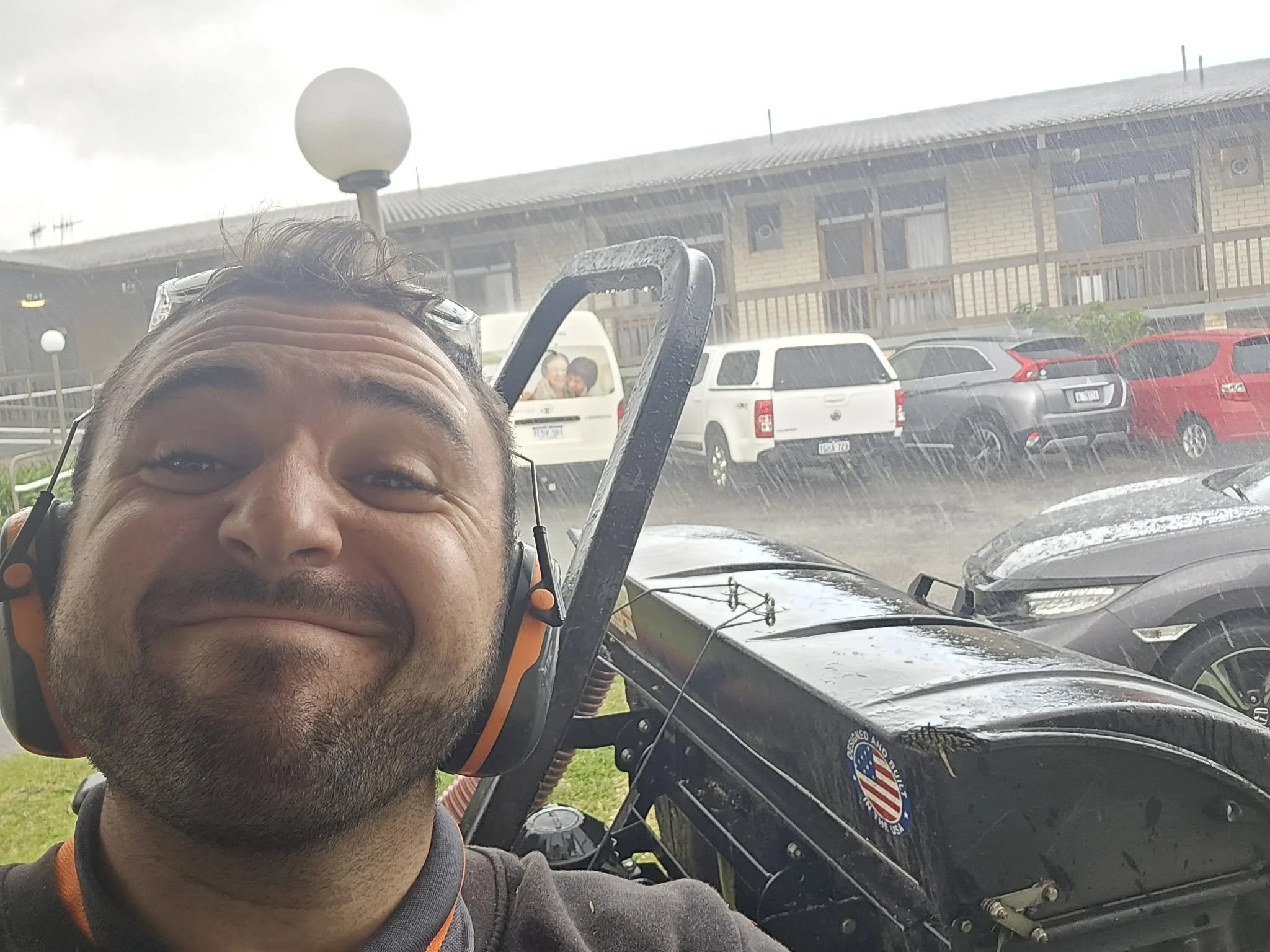 A man with a beard and glasses resting on his head taking a selfie in the rain, with a utility vehicle behind him, and a parking lot with parked cars and a building in the background.