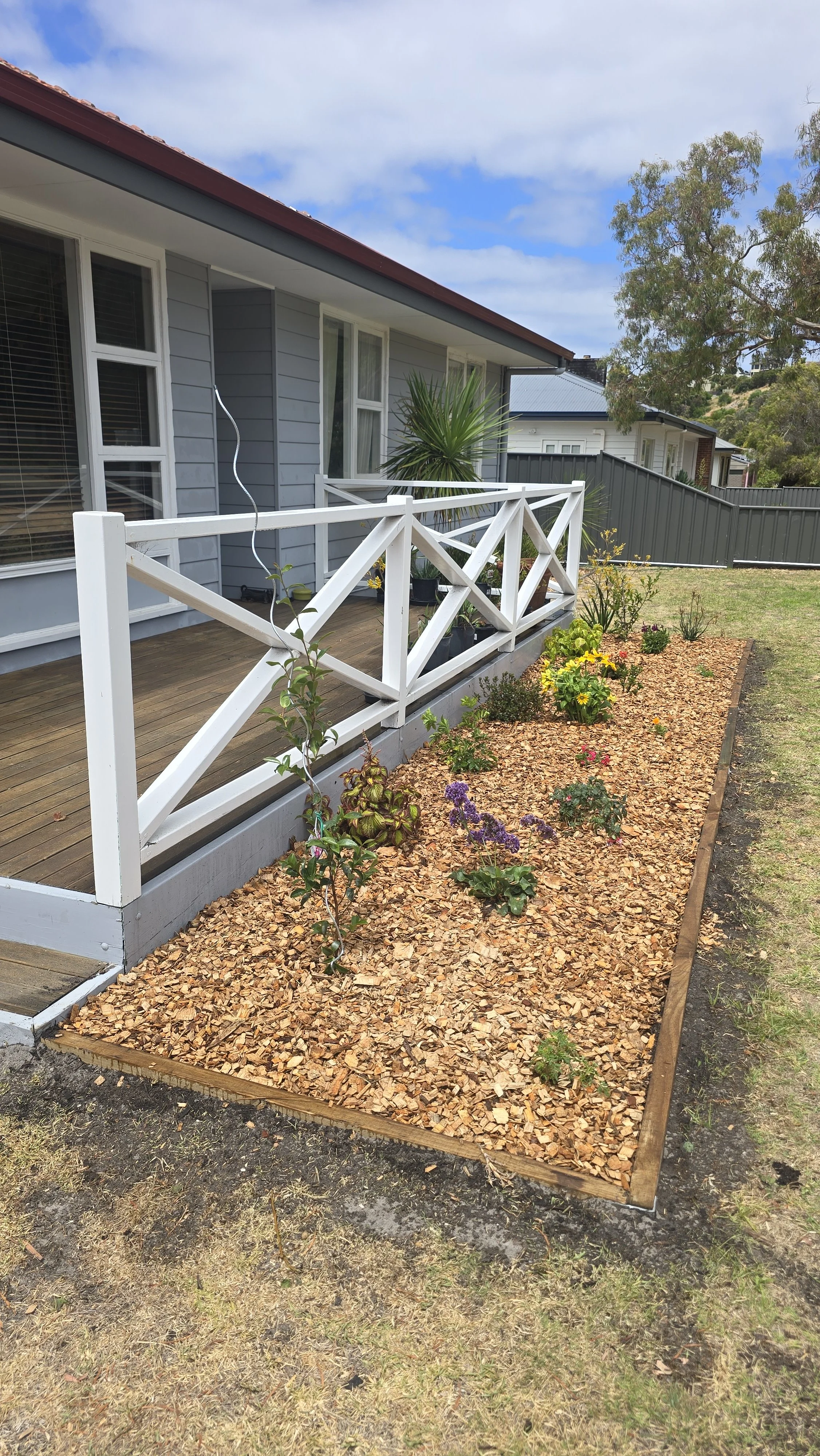 A small front garden with a wooden border, covered with wood chips, and planted with various small flowers. A wooden deck with a white railing extends from a gray house with large windows. A large potted plant with long, pointed leaves is on the deck. A wire is hanging from the railing. The background features a cloudy sky and neighboring houses with fences.