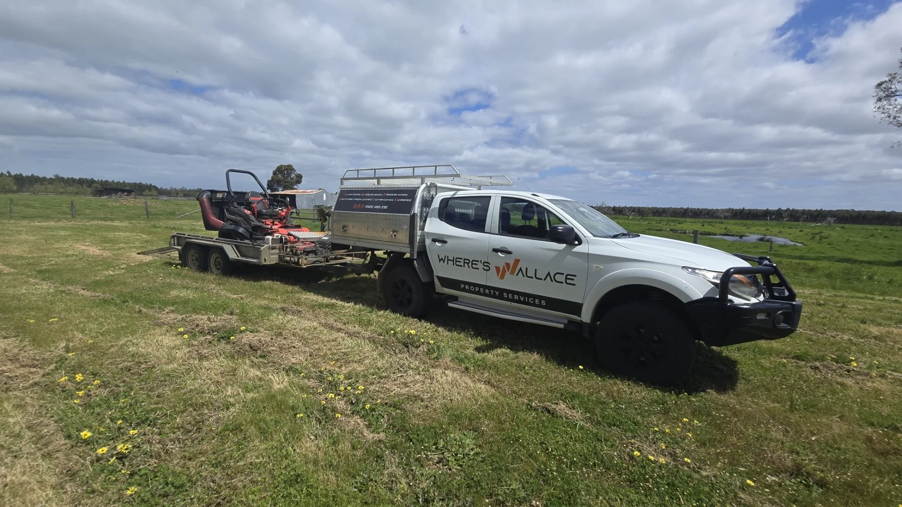 A white pickup truck with company branding is parked on a grassy field, towing a trailer with a red lawn mower in a rural area under a cloudy sky.
