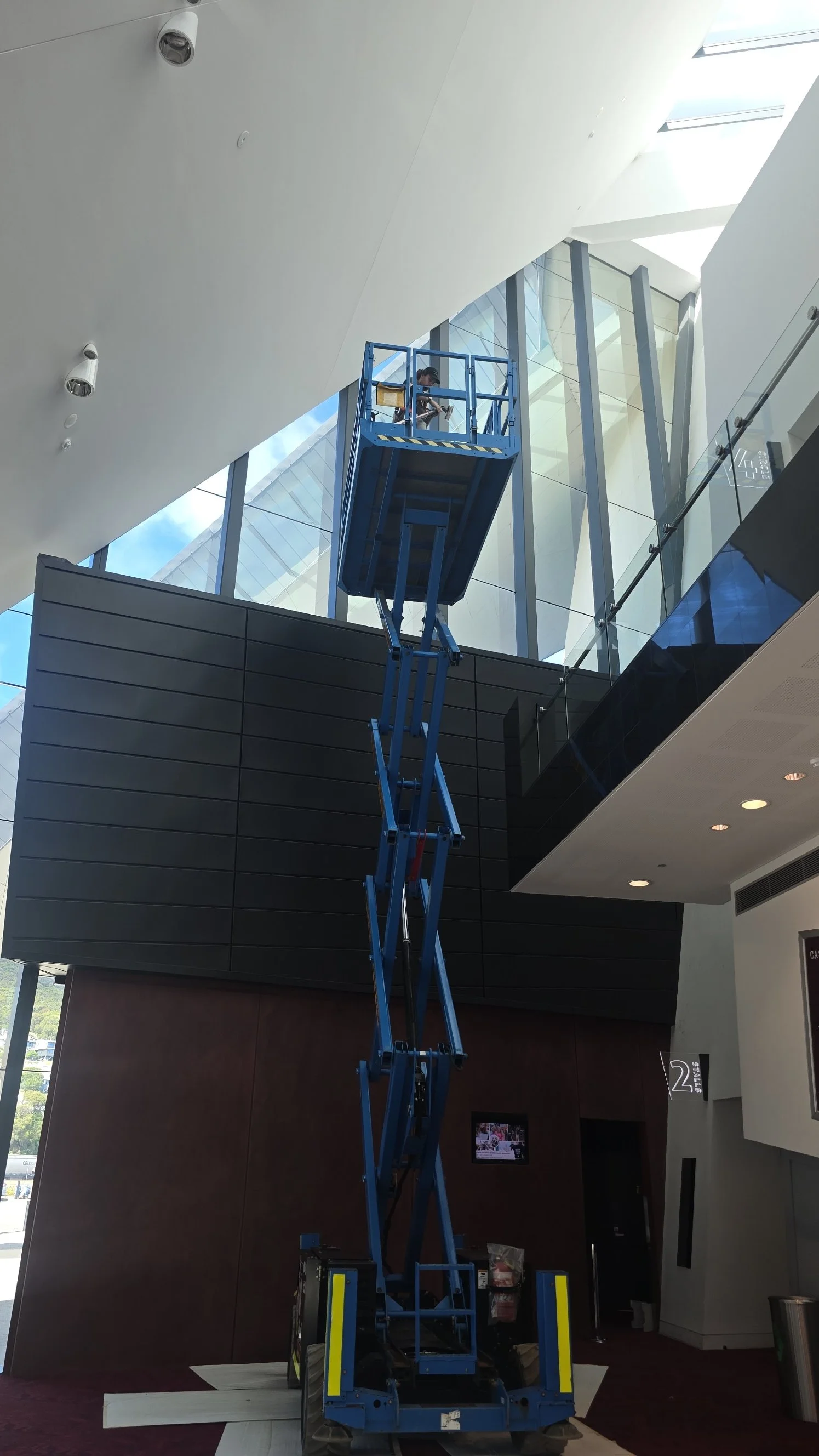 A person working on a blue scissor lift inside a modern building with large windows and high ceilings.