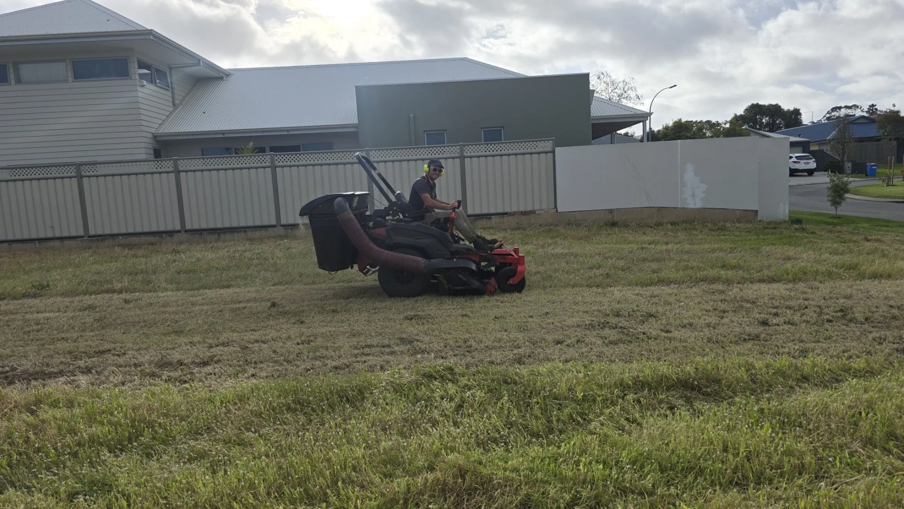 A man with headphones riding a riding lawn mower on a grassy field in a residential neighborhood with houses, a fence, trees, and a cloudy sky in the background.