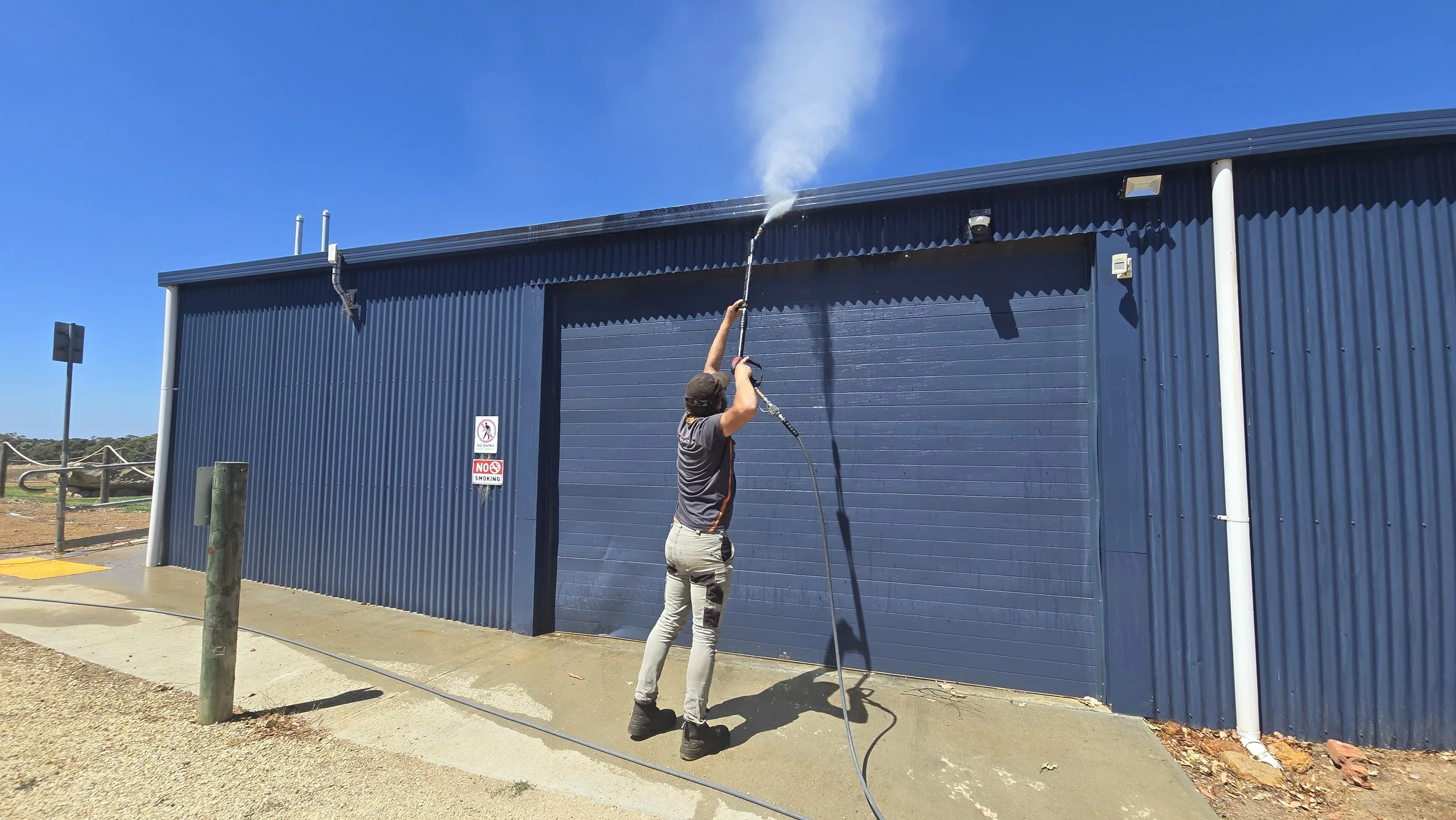 A person is pressure washing the exterior blue metal wall of a building on a sunny day.