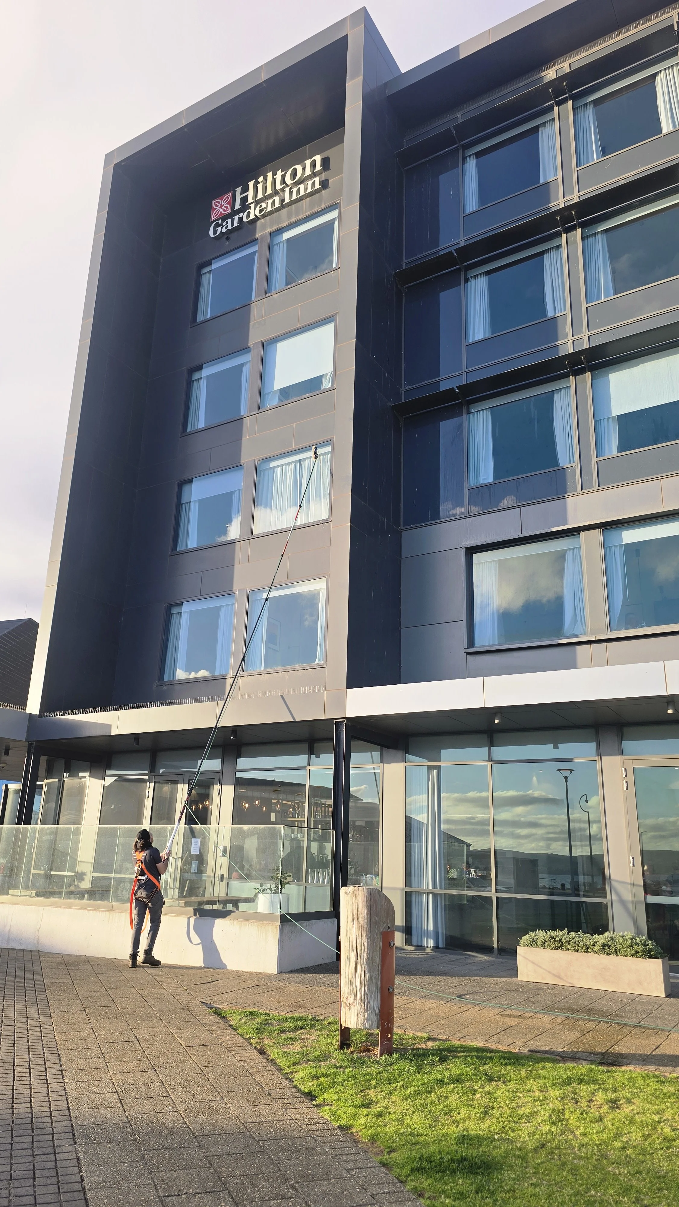 The image shows a modern multi-story hotel building with a sign that reads 'Hilton Garden Inn' near the top. In front of the hotel, a person is using a cleaning tool connected to a long pole, reaching up to the top of the building. The building has large windows with curtains, and the bottom floor appears to have a lobby area with glass doors and windows. The surrounding area includes a paved sidewalk, some green grass, and a few street lamps.
