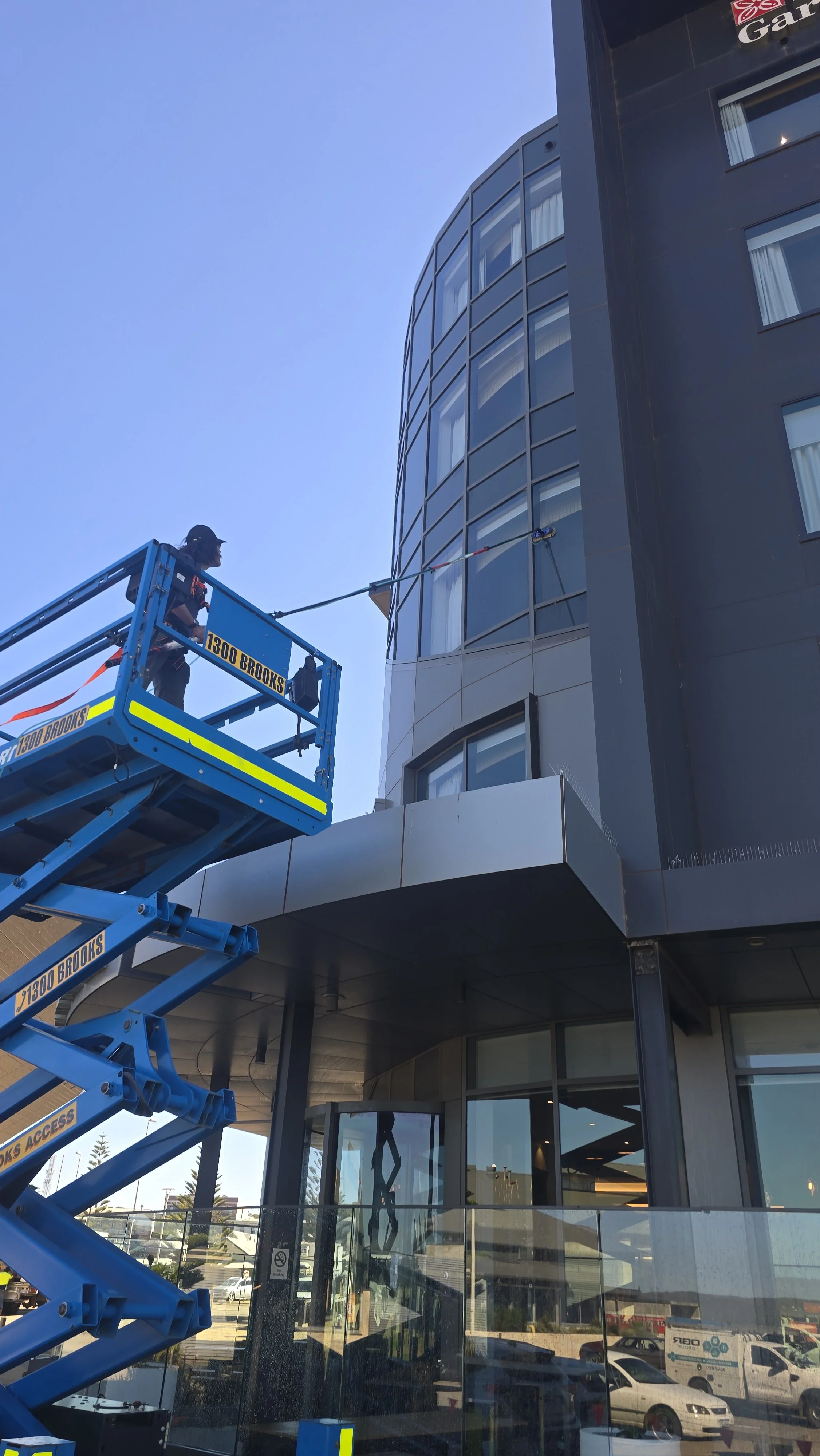 A worker on a blue hydraulic lift cleaning the exterior windows of a tall modern building with a curved glass facade and dark exterior panels.