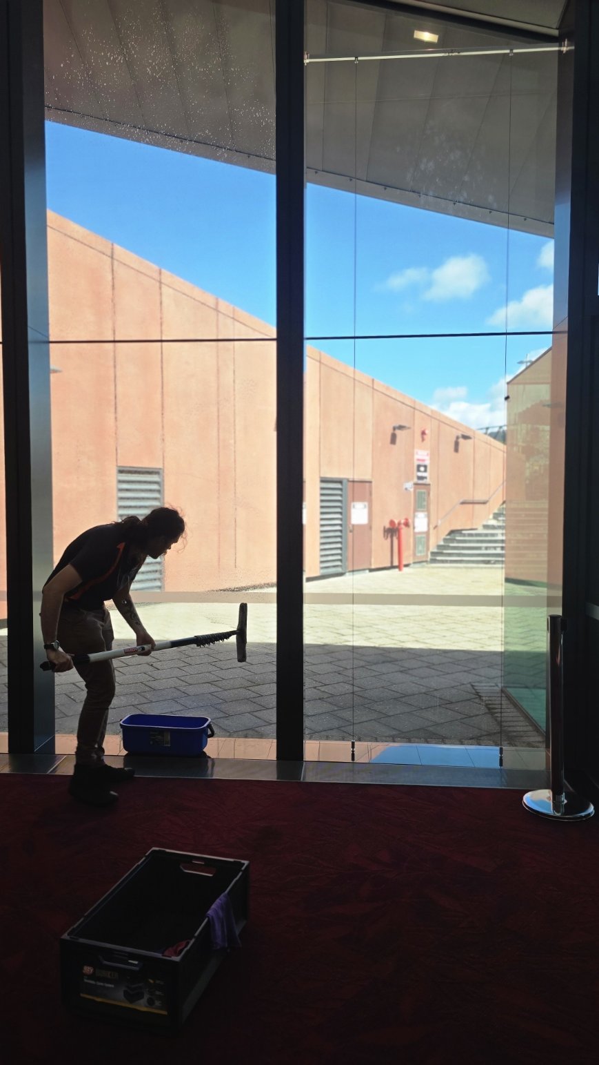 A woman cleaning the floor in a building lobby with a squeegee. Outside, a brick building and blue sky are visible through large glass windows.