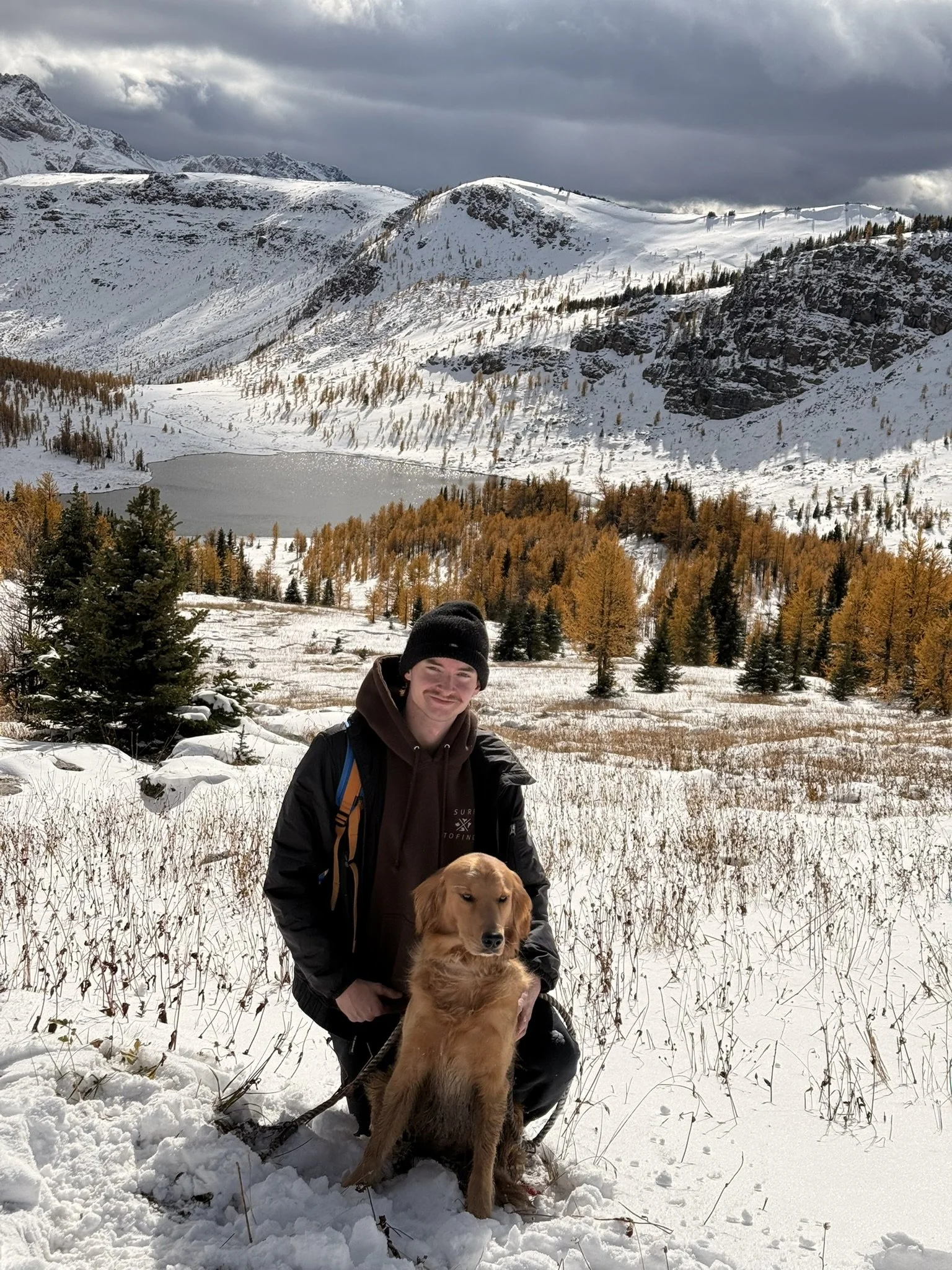 A person wearing a black beanie and black jacket kneeling in the snow with a golden retriever in an mountain landscape. Snow-covered mountains, a lake, and autumn-colored trees are in the background, with a cloudy sky overhead.
