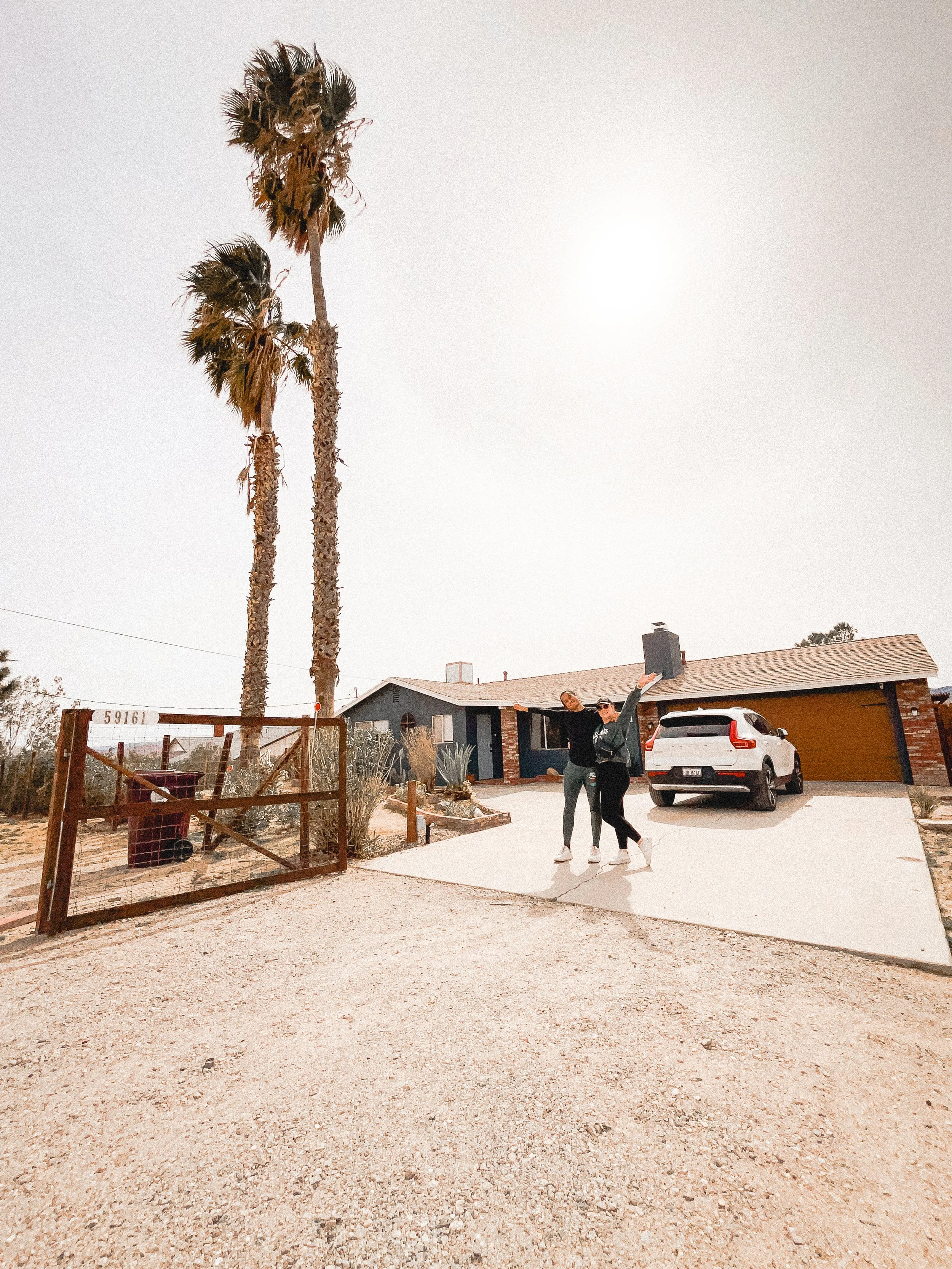 Two women standing in front of a house with a white car parked in the driveway, palm trees nearby, during daytime.