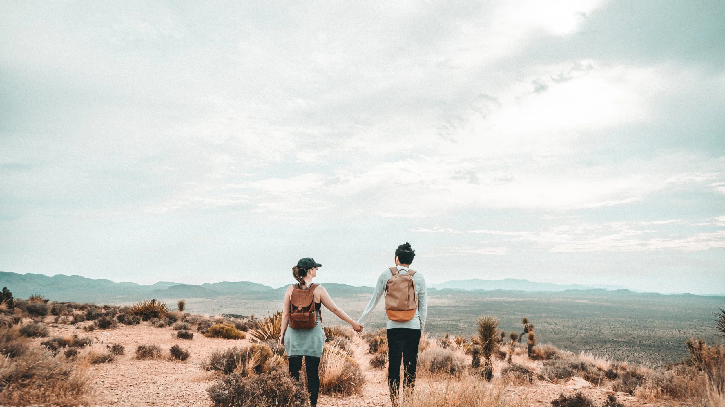 Two people hiking in a desert landscape with mountains in the distance, holding hands as they walk along a trail under a cloudy sky.