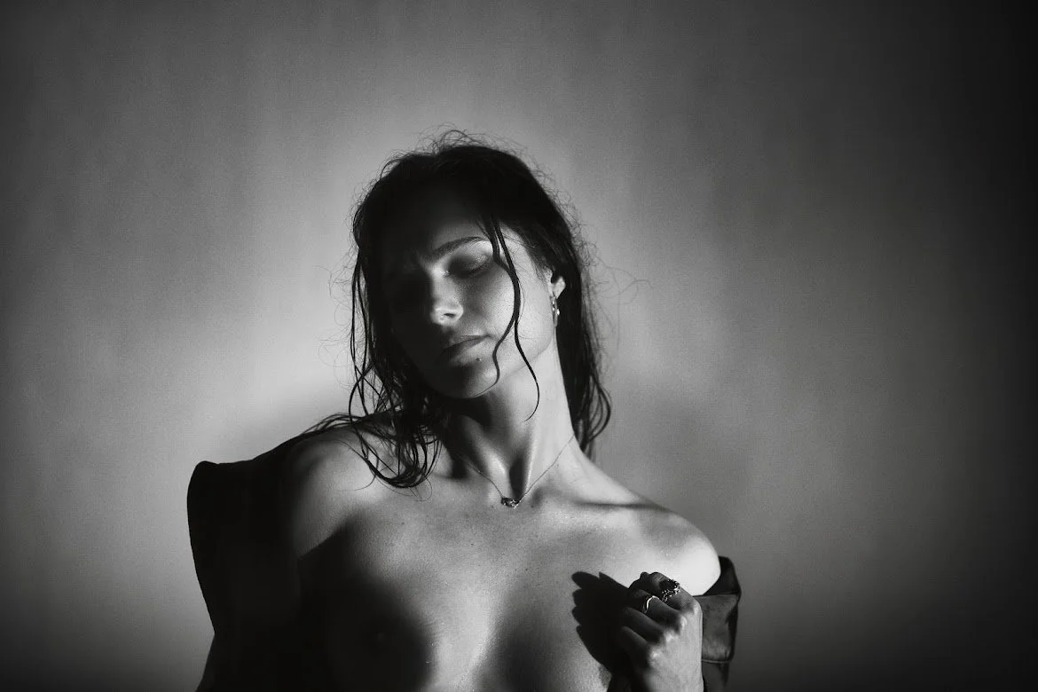 Black and white photo of a woman with wet hair, topless, embracing a dark garment near her shoulder, with soft shadows and a plain background.