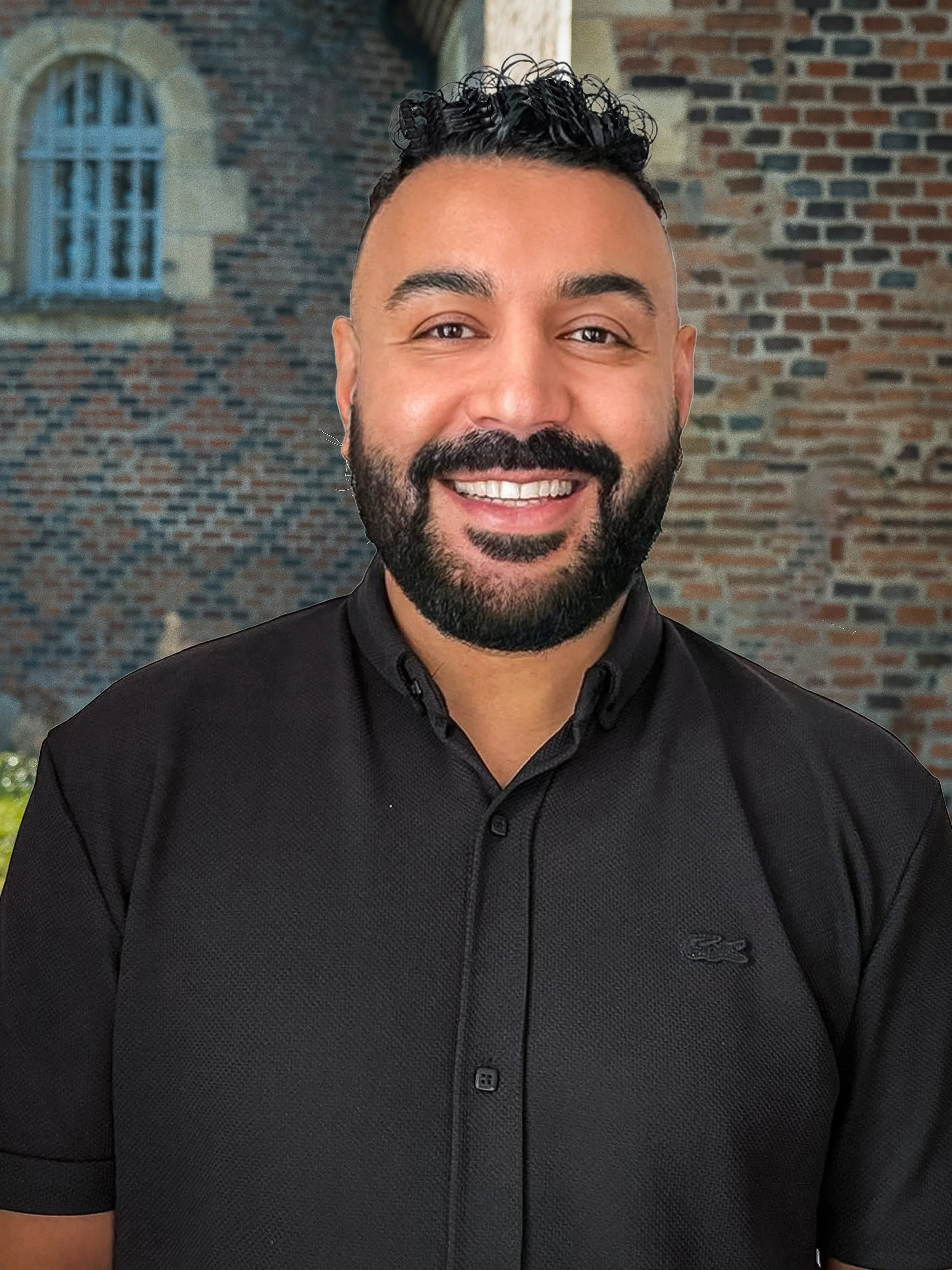 Homme souriant avec barbe et cheveux courts bouclés, portant une chemise noire, en arrière-plan un mur en briques avec une fenêtre.