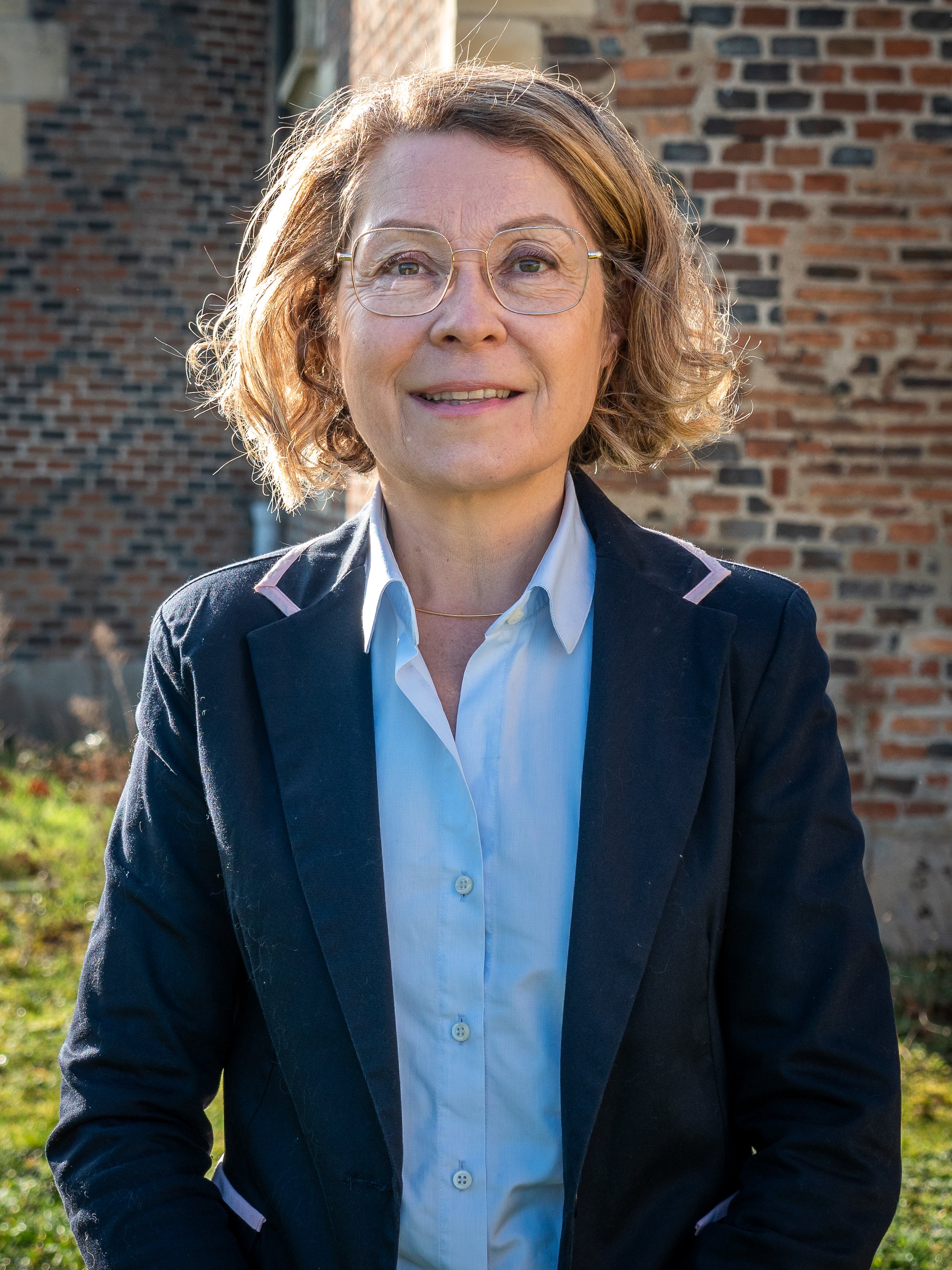 Une femme d'âge moyen avec des lunettes, portant une chemise blanche et une veste noire, debout devant un mur en briques, dans un endroit en extérieur