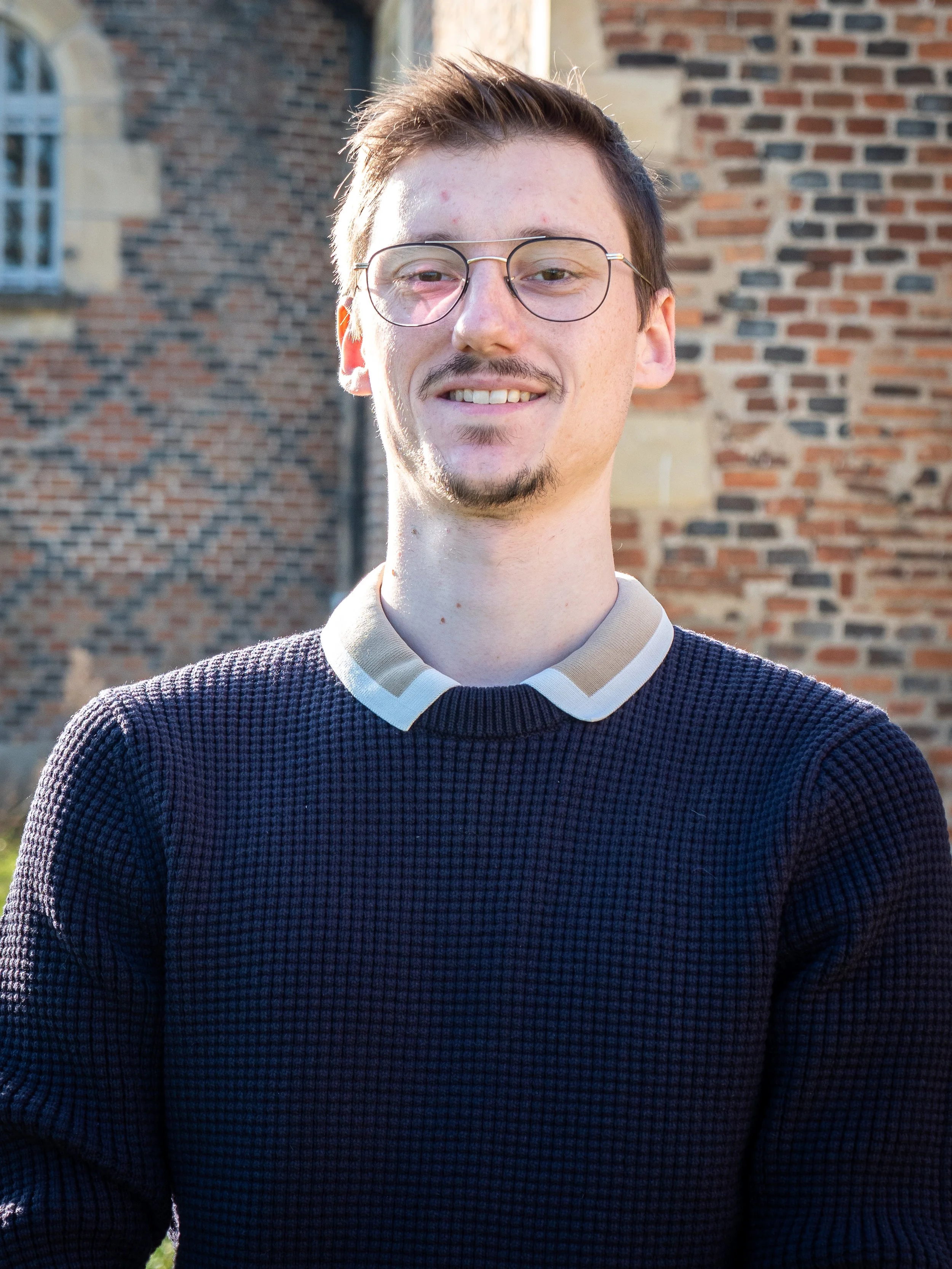 Jeune homme avec lunettes, souriant, portant un pull noir avec un col beige, devant un mur en briques.