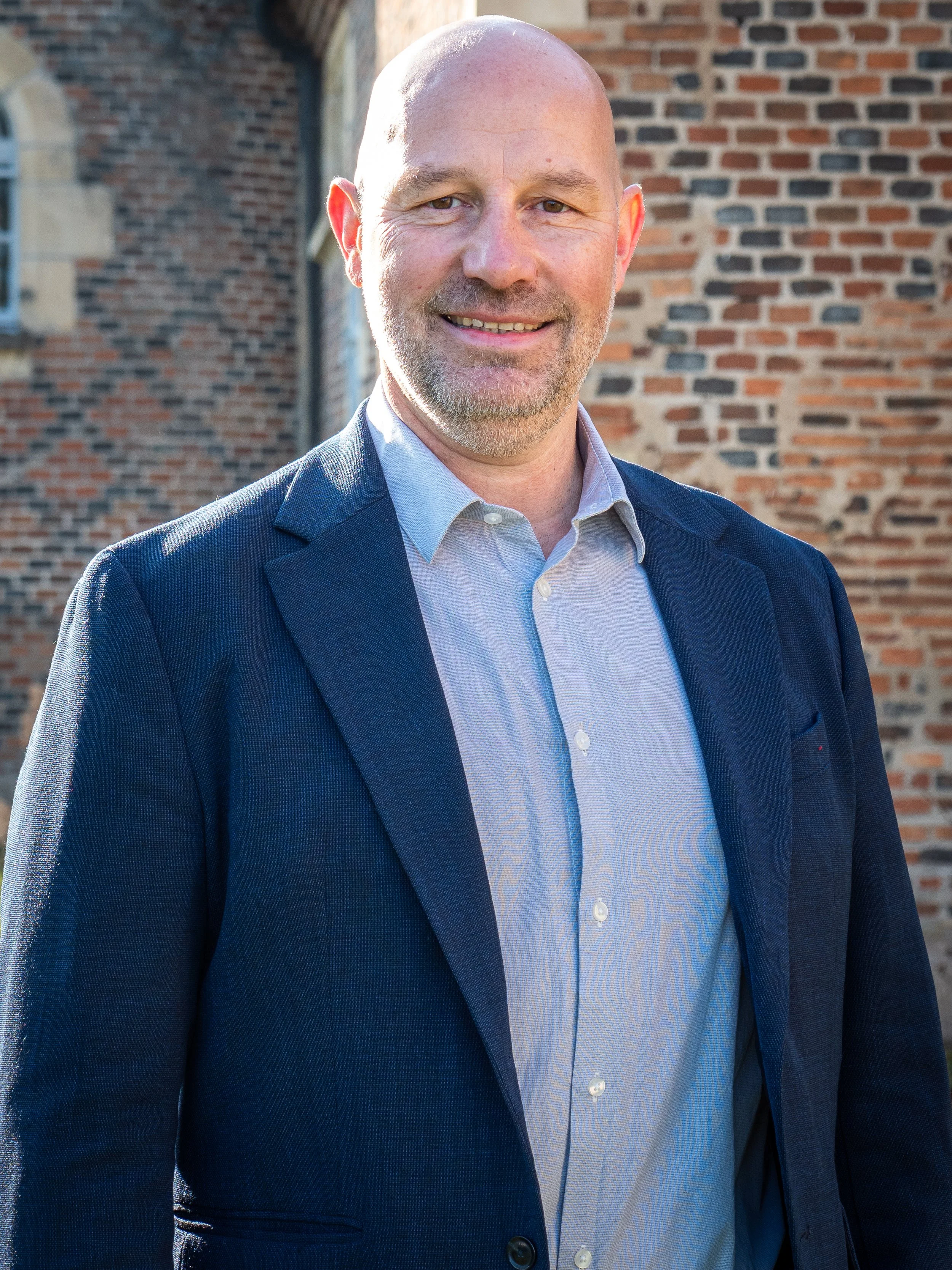 Hommes souriant portant un costume bleu clair et une chemise blanche, debout devant un mur en briques.