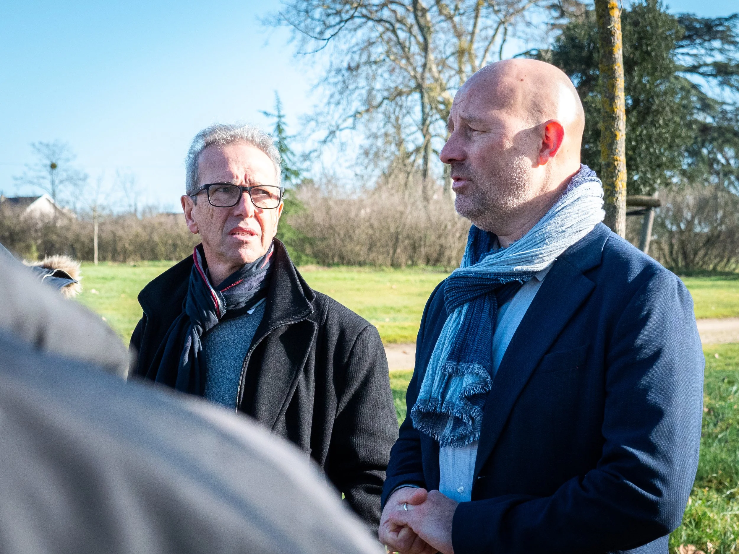 Deux hommes en discussion en plein air, une scène ensoleillée avec des arbres et un champ en arrière-plan.
