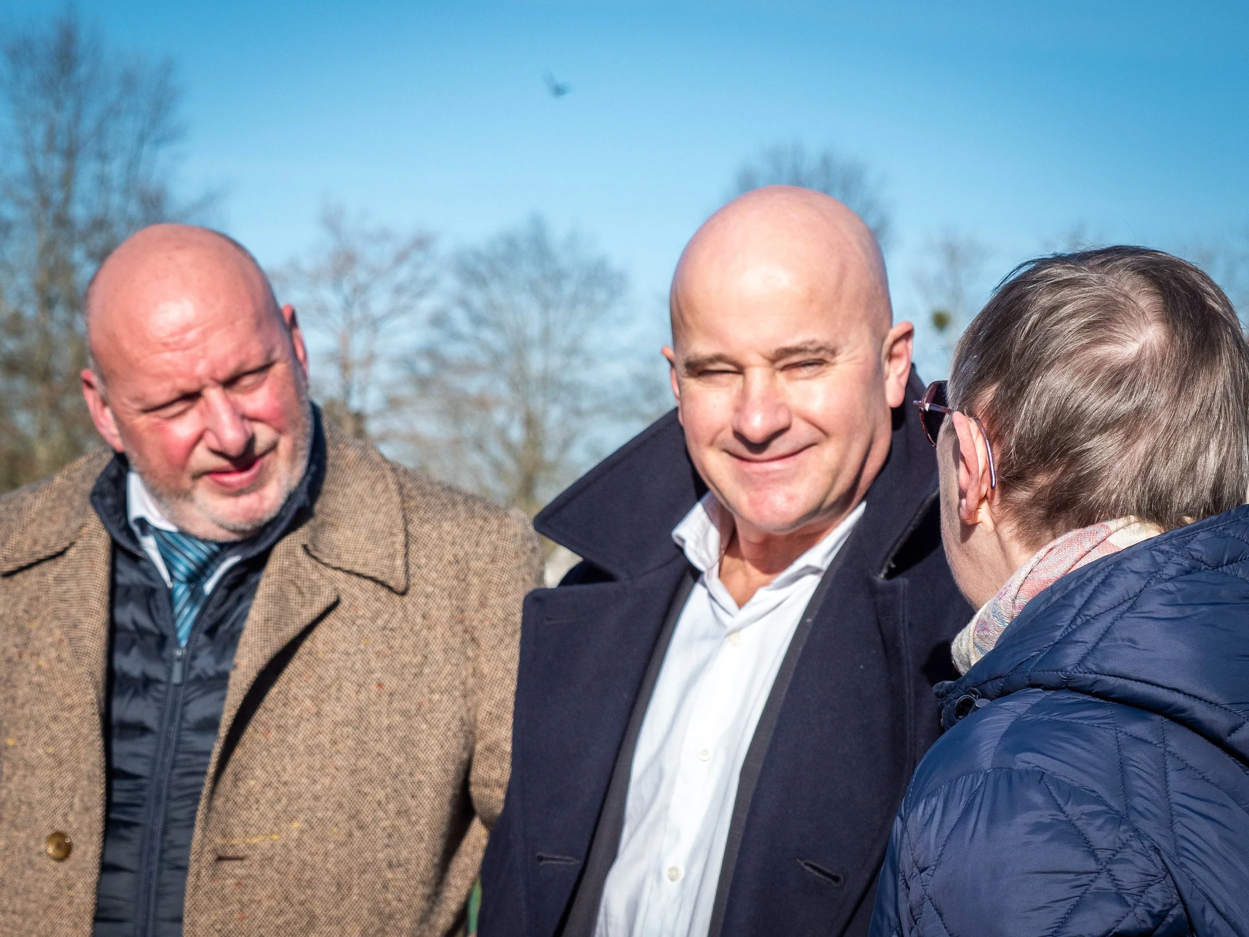 Trois personnes parlent en extérieur sous un ciel bleu, deux hommes et une femme, tous portant des manteaux.