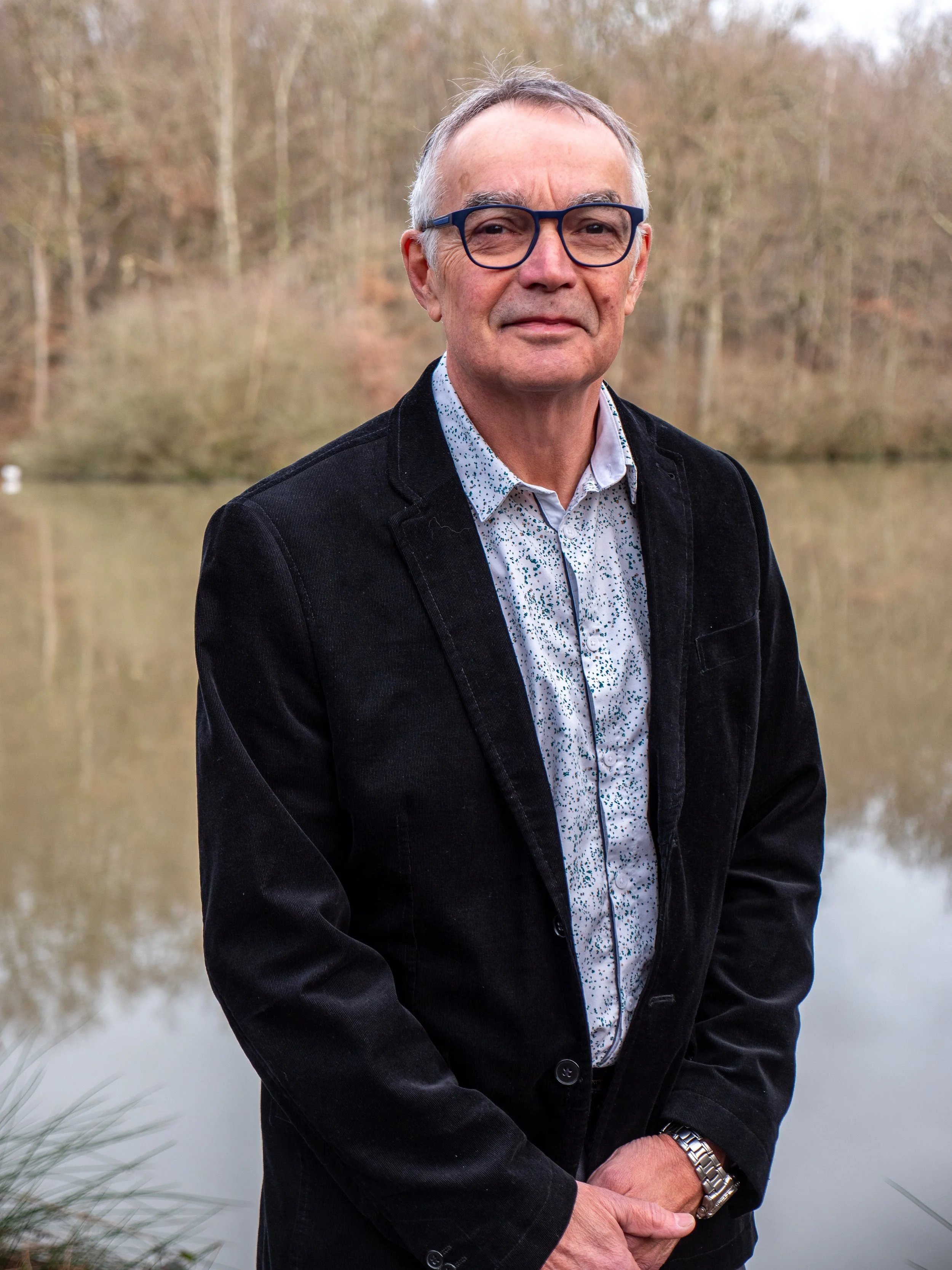 Un homme âgé portant des lunettes, une chemise à motifs, et une veste noire, se tient devant un lac avec des arbres en arrière-plan.