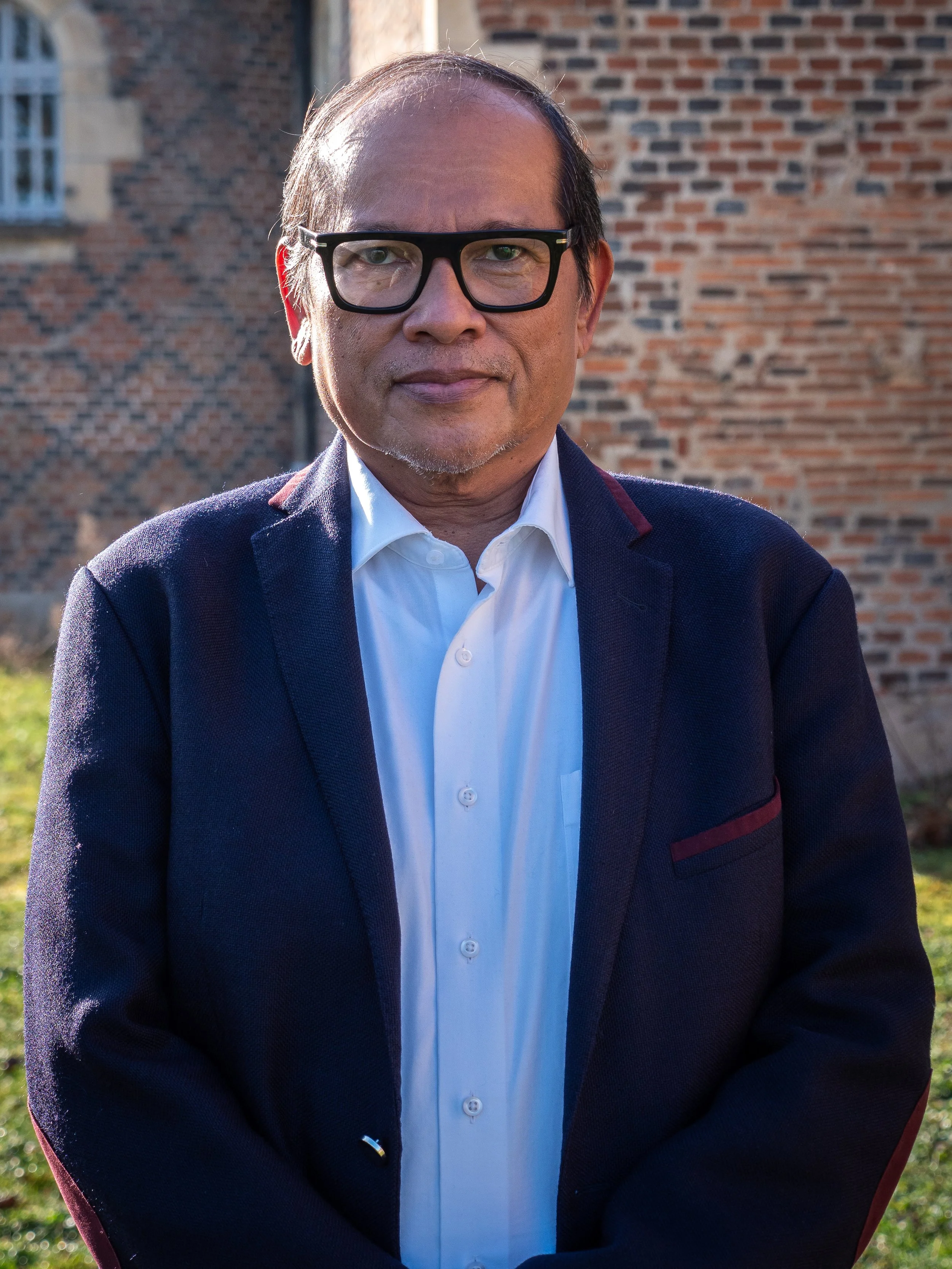 Homme portant des lunettes noires, chemise blanche et veste sombre, debout à l'extérieur devant un mur en briques rouges.
