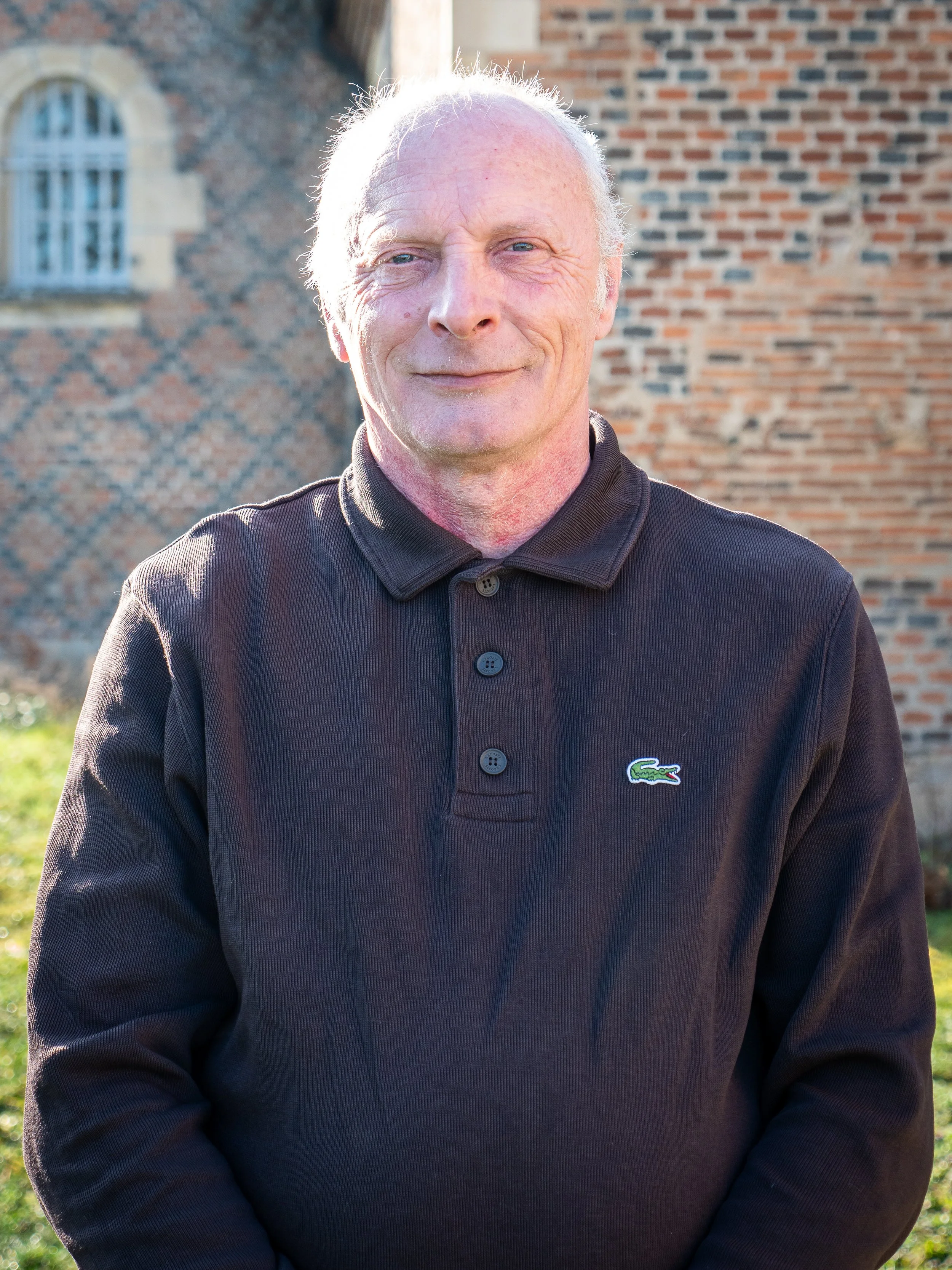 Un homme âgé souriant, vêtu d'un polo Lacoste noir, debout devant un bâtiment en briques, en extérieur par une journée ensoleillée.
