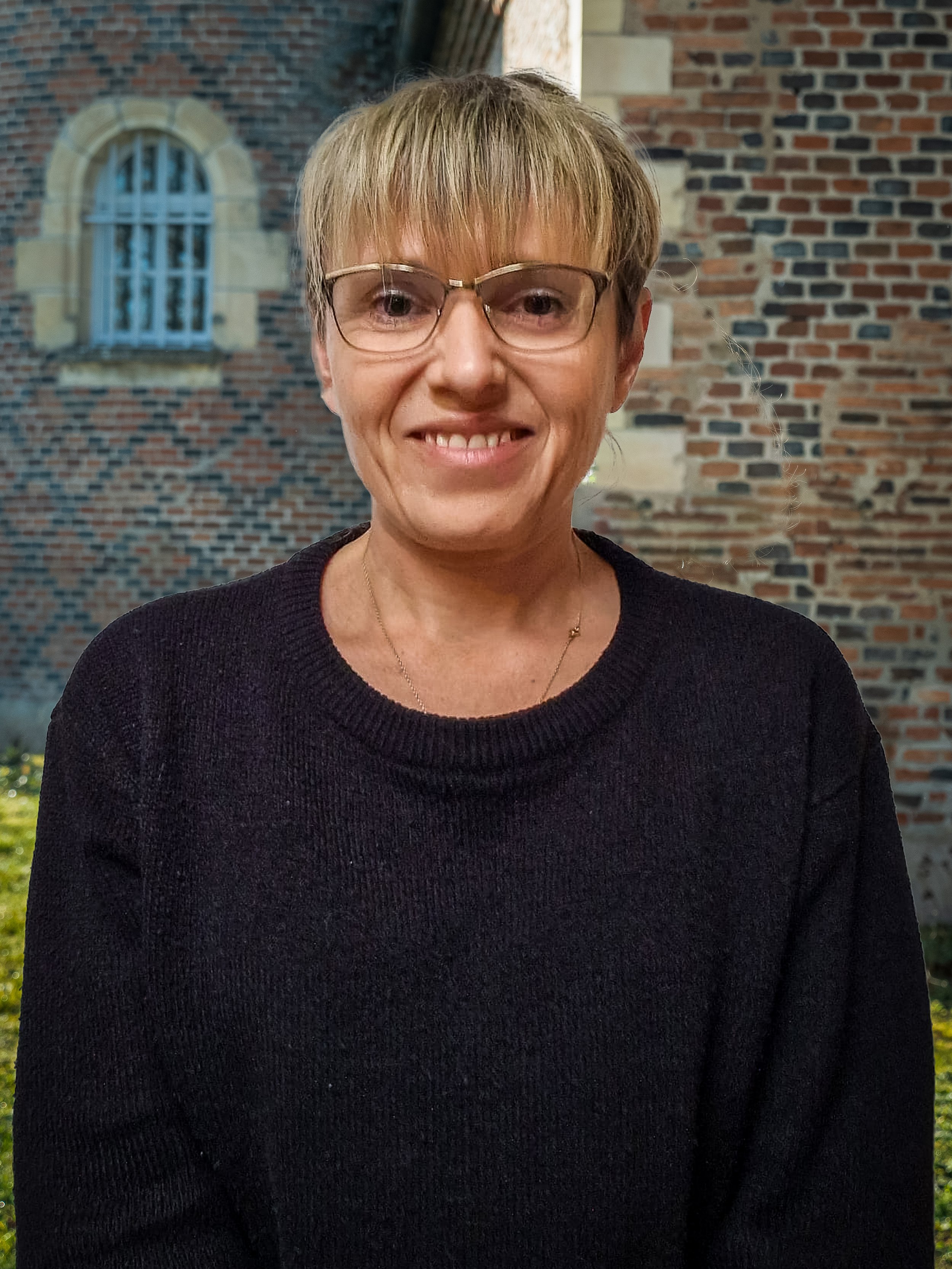 Une femme souriante portant des lunettes, vêtue d'un pull noir, devant un mur en briques avec une fenêtre.