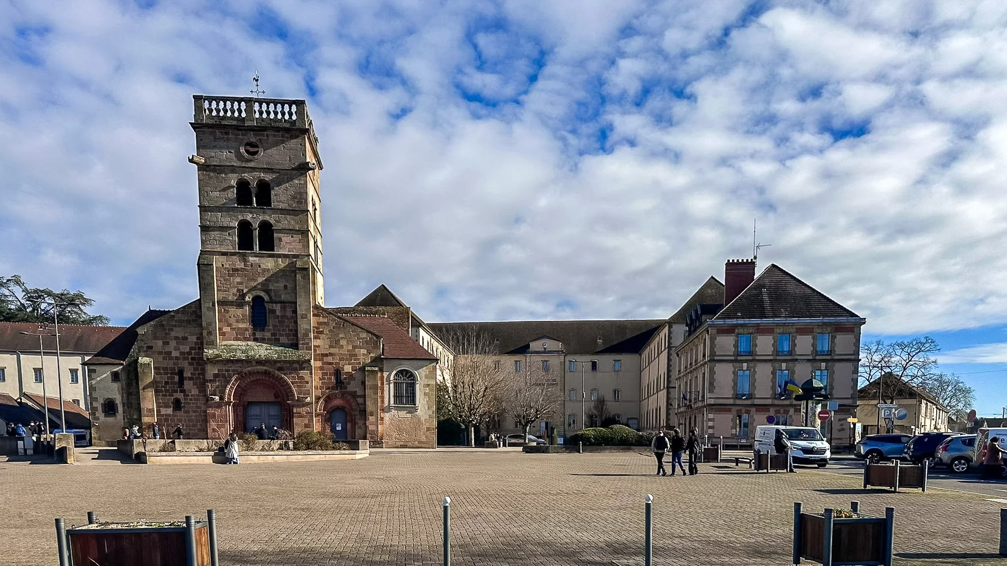 Une place publique avec une ancienne bâtisse en pierre avec une tour, entourée de bâtiments résidentiels, dans une journée ensoleillée avec un ciel partiellement nuageux.