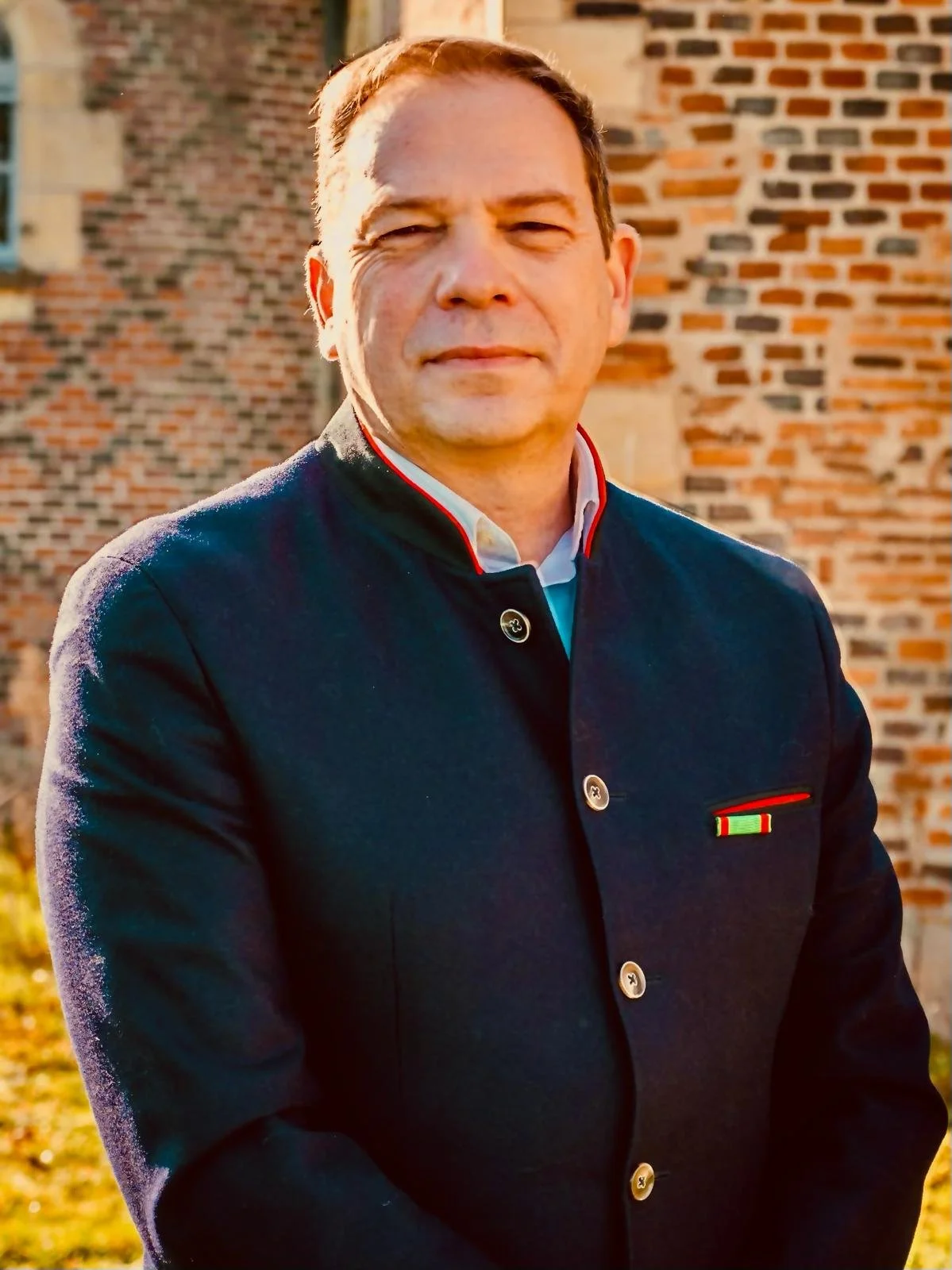 Un homme en uniforme militaire avec une veste sombre, portant une médaille, se tient devant un mur en briques, en plein air, sous la lumière du soleil.
