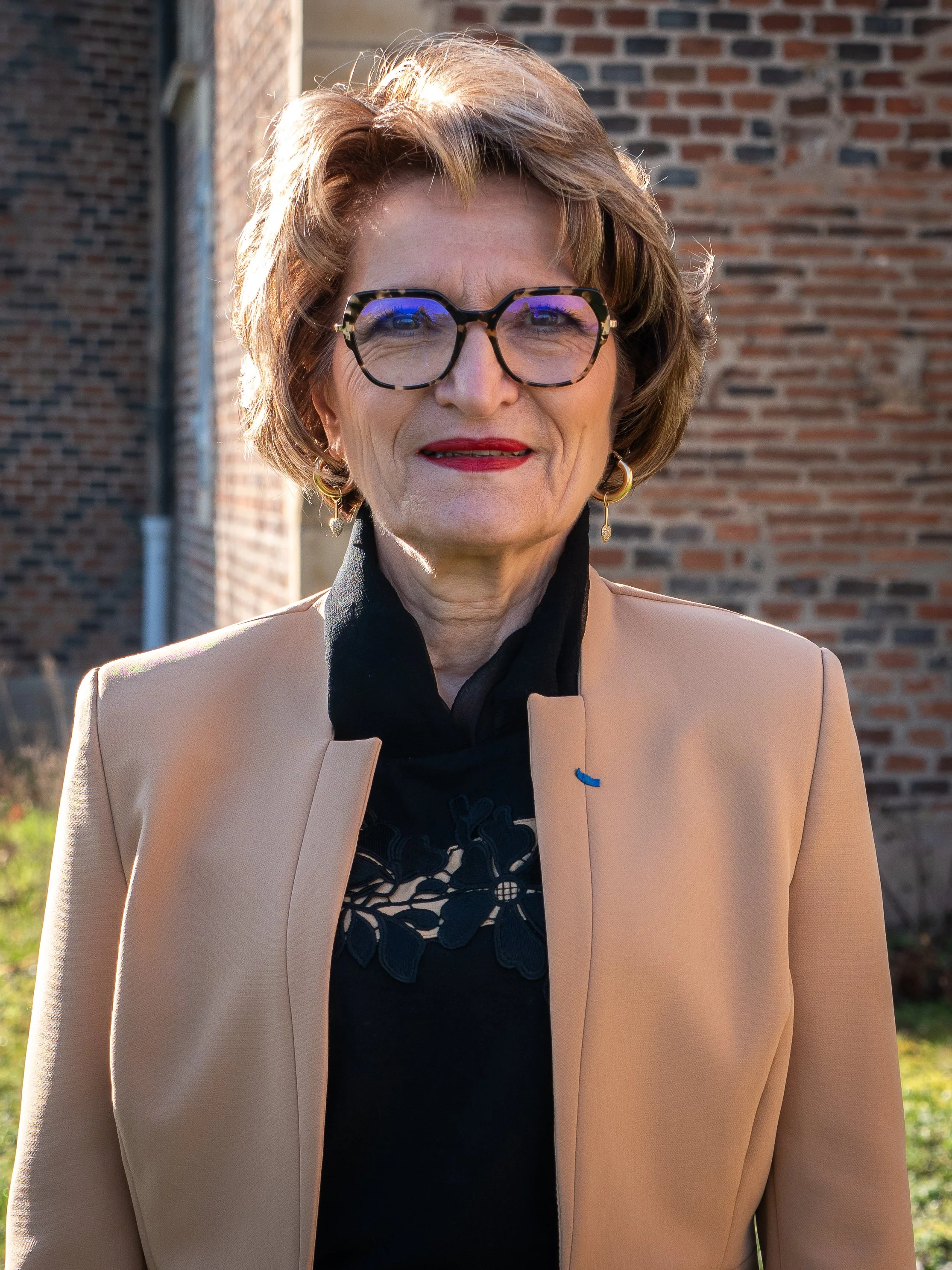 Femme âgée portant des lunettes à monture tachetée, une veste beige, un haut noir avec broderie florale, et des boucles d'oreilles en forme de goutte, dehors près d'une maison en briques.