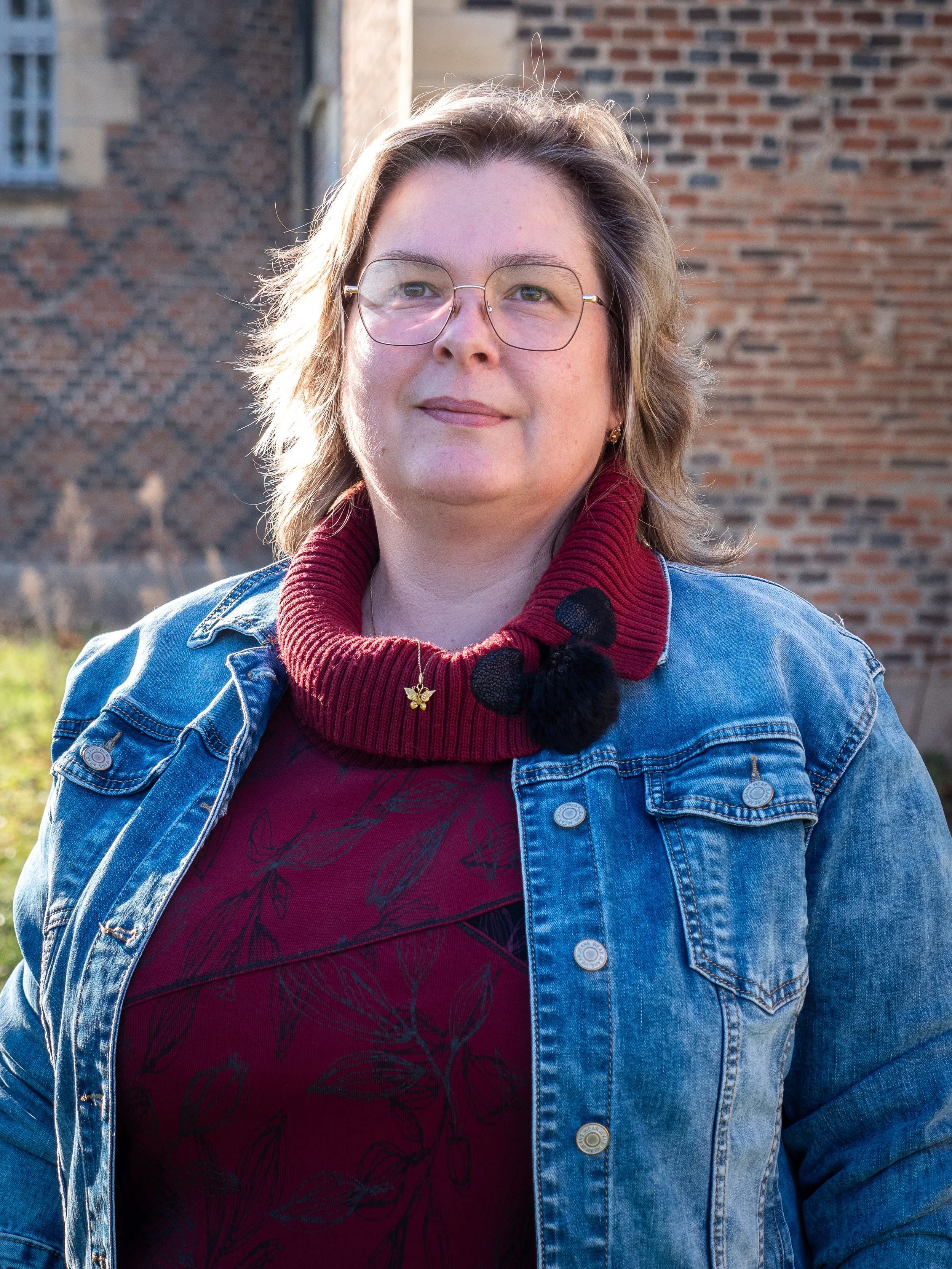 Une femme porte des lunettes, un pull rouge avec un col roulé, une veste en jean et un collier avec un pendentif en forme de papillon. Elle se tient devant un mur en briques, dans un endroit en plein air, ensoleillé.
