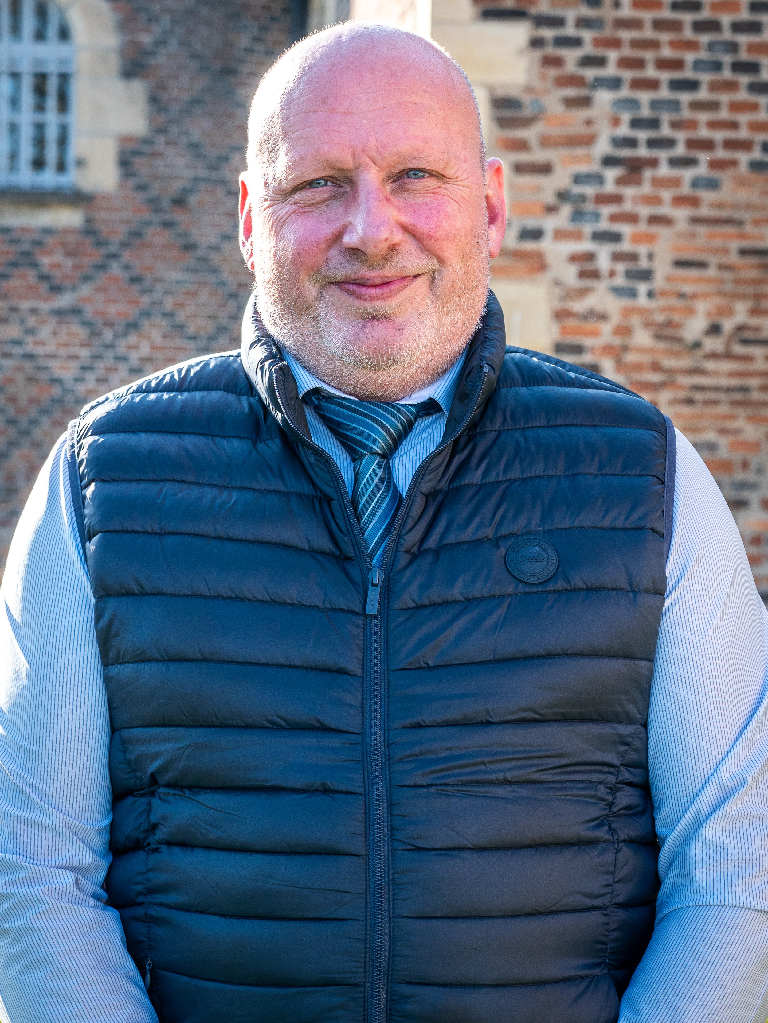 Portrait d'un homme souriant, portant une veste à puffer noire, une chemise bleue à rayures et une cravate, devant un bâtiment en briques.