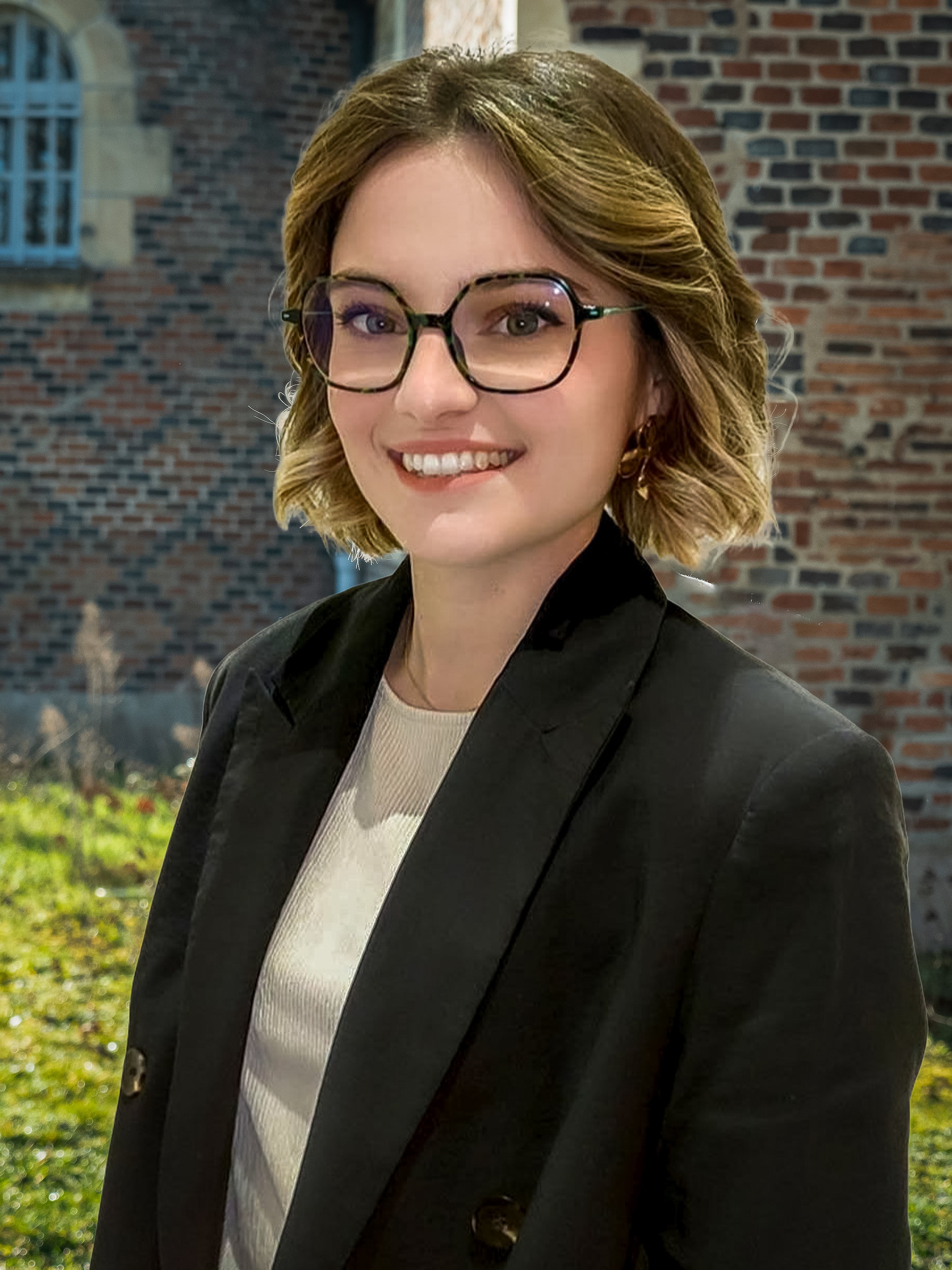Jeune femme aux cheveux bouclés, portant des lunettes et un blazer noir, souriant devant un mur en briques.