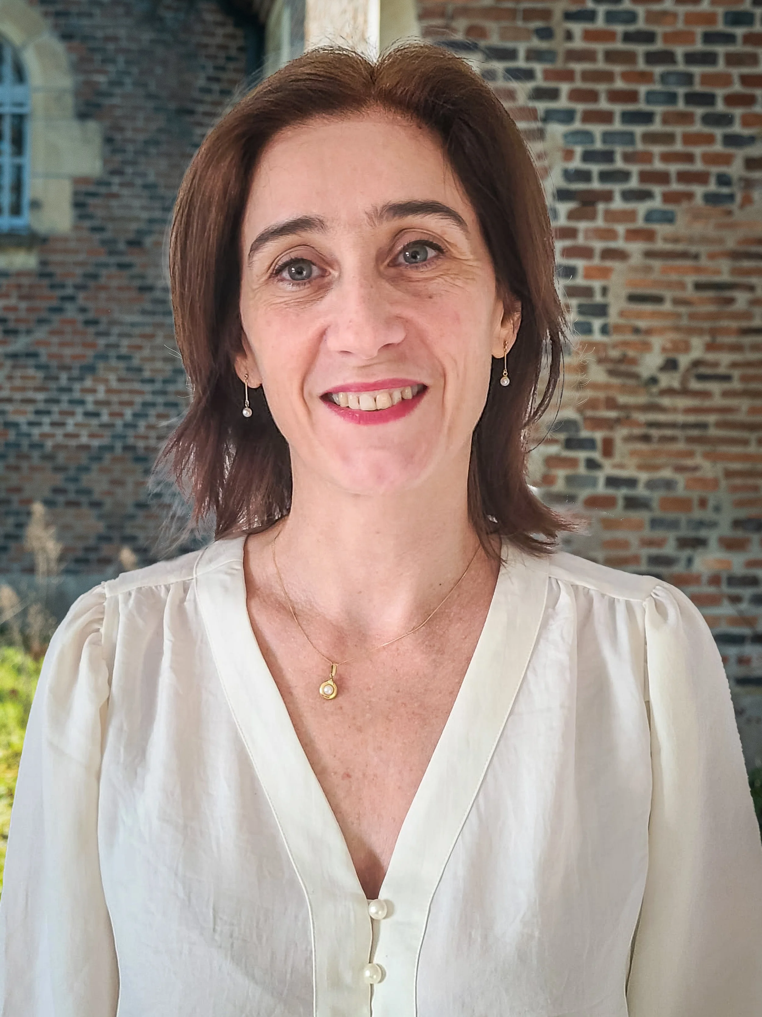 Portrait d'une femme souriante portant une chemise blanche, avec un collier et des boucles d'oreilles, devant un mur en briques.