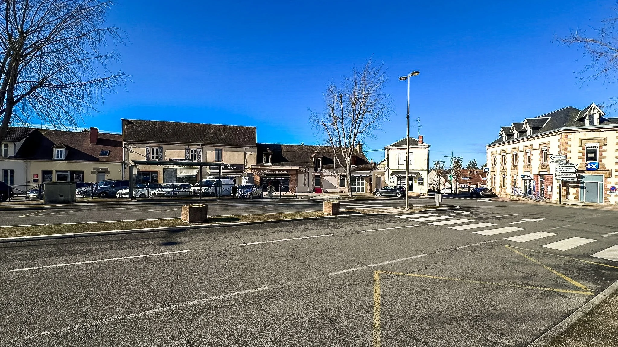 Une place de village avec un parking, des bâtiments résidentiels et commerciaux, un arbre sans feuilles, un ciel bleu clair, un passage piéton, et une rue avec quelques voitures
