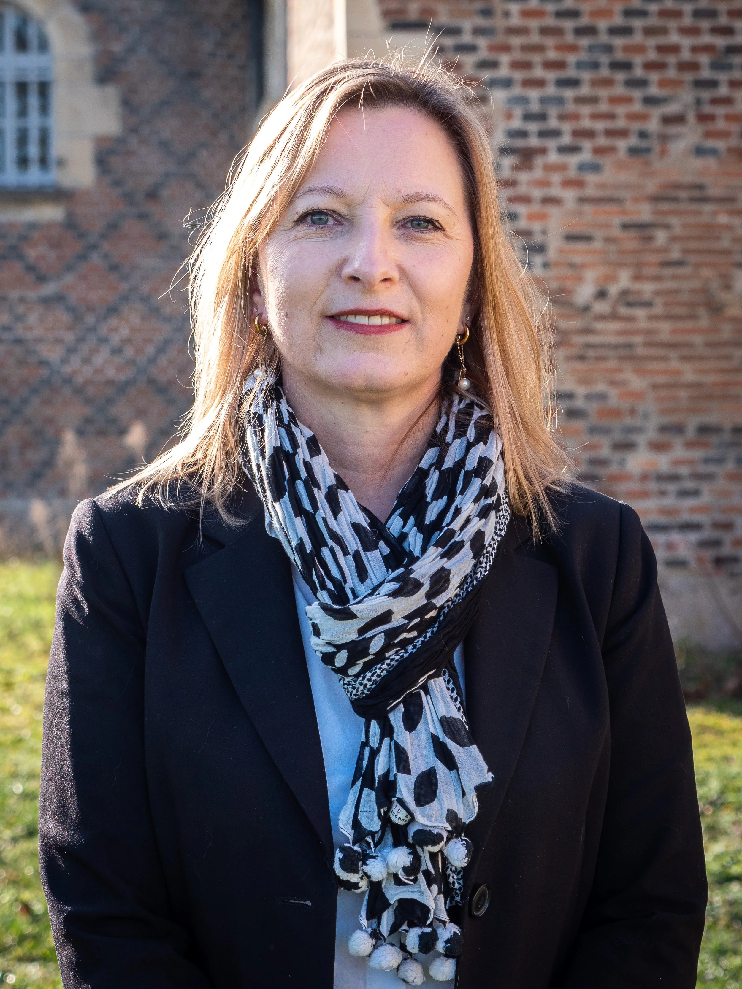 Une femme souriante avec des cheveux blonds, portant un manteau noir, une chemise blanche et une écharpe à motifs noir et blanc, se tenant à l’extérieur avec un bâtiment en brique en arrière-plan.