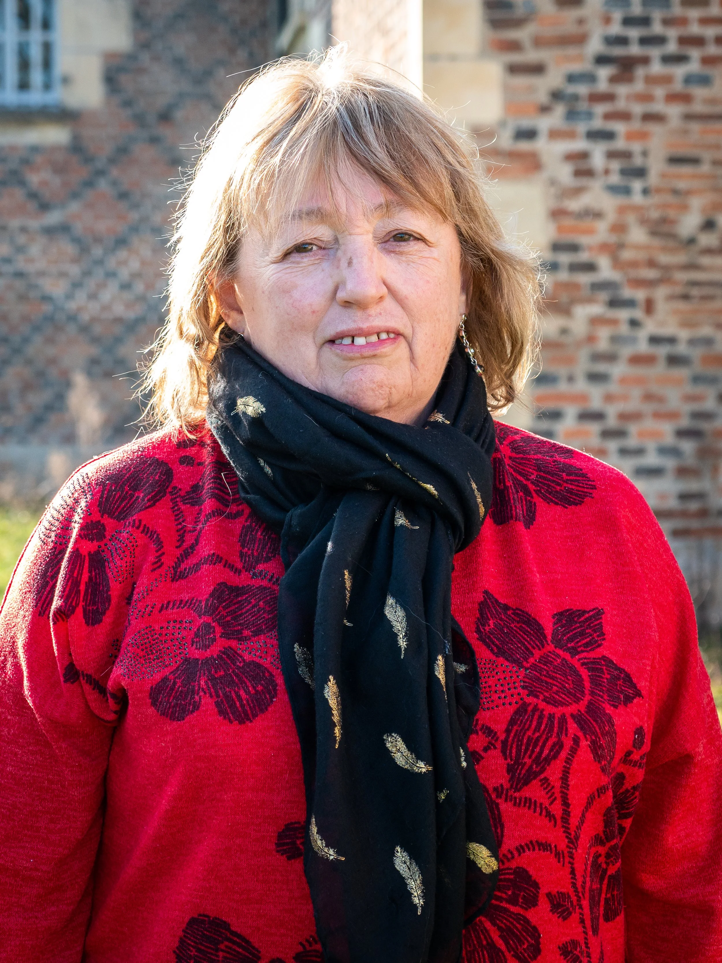 Une femme âgée portant un pull rouge avec un motif floral noir et une écharpe noire avec des plumes dorées. Elle se tient devant un bâtiment en briques.