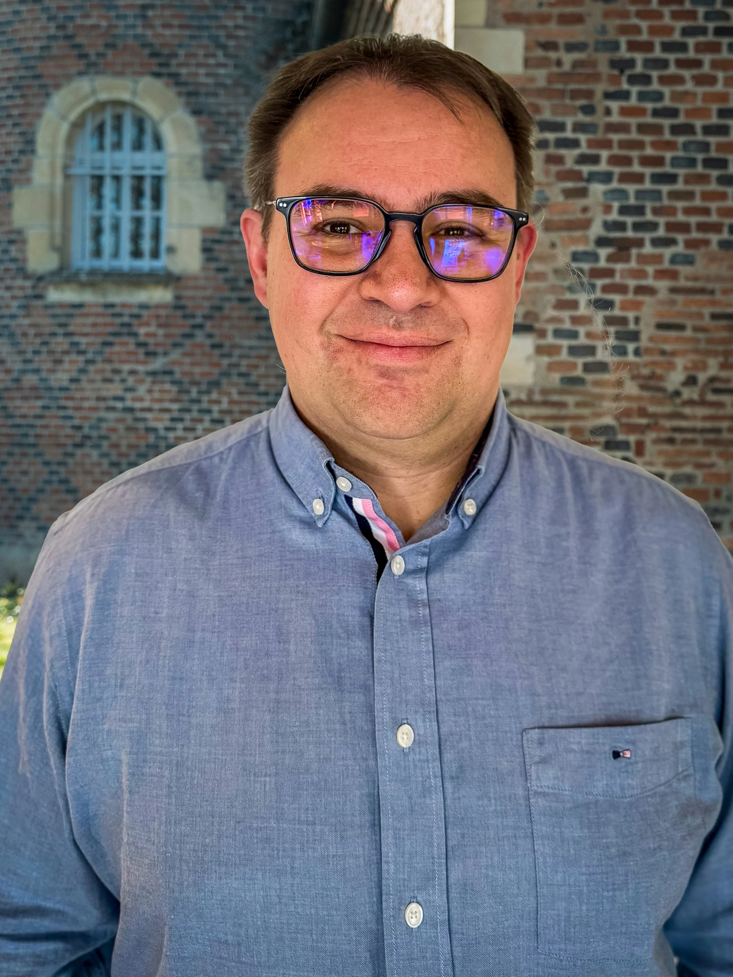 Portrait d'un homme souriant portant des lunettes, en chemise bleue, avec un mur de briques en arrière-plan.