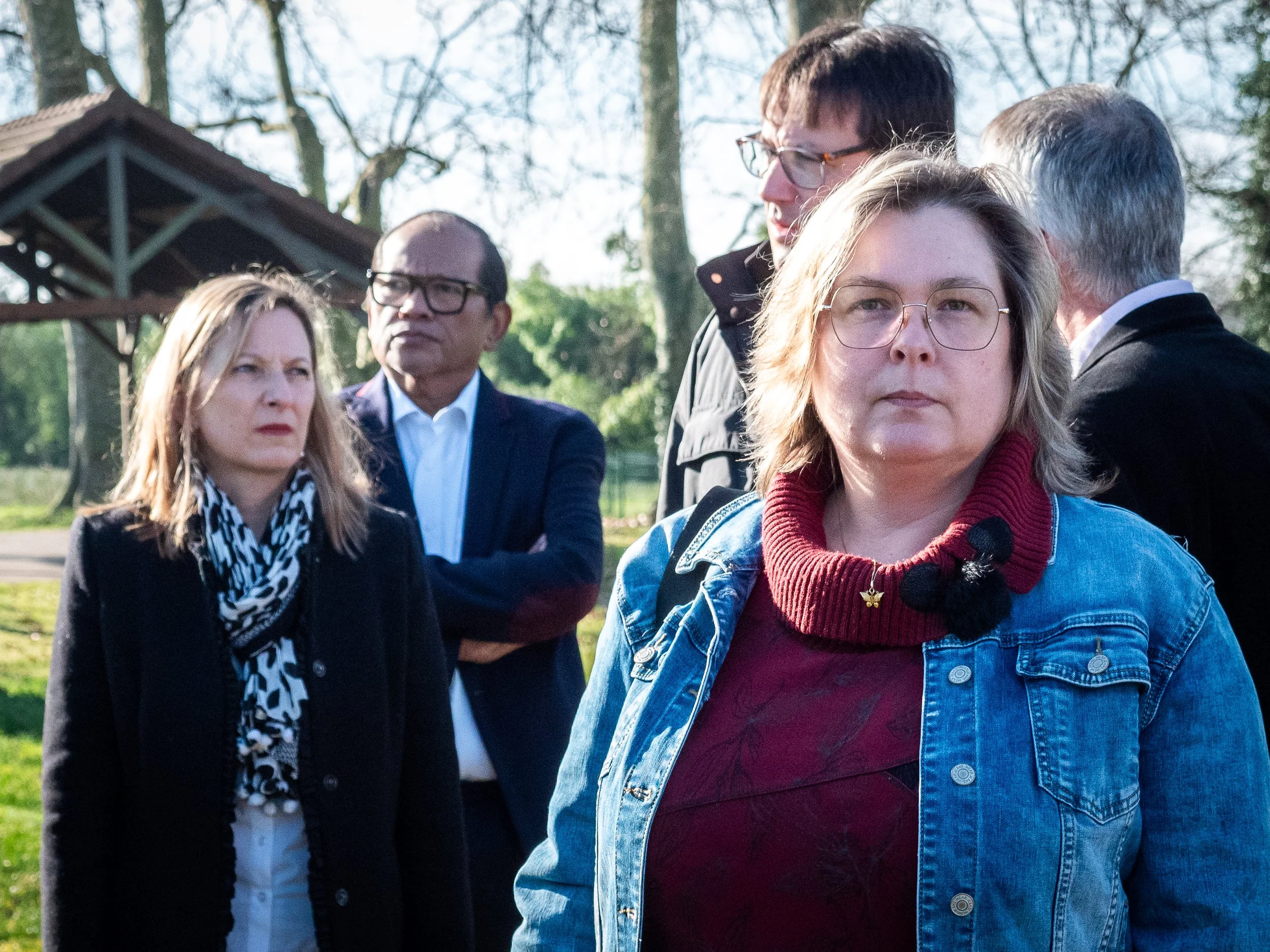 Groupe de cinq personnes, dont deux femmes et trois hommes, en extérieur près d'arbres et d'une petite structure en bois, avec expression sérieuse.