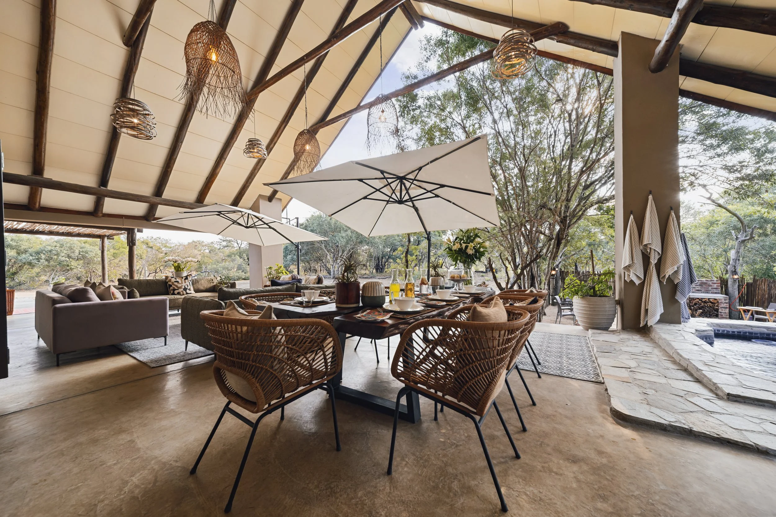 Open-air patio with a wooden ceiling, hanging wicker lights, large white umbrellas, a dining table set with breakfast items, and patio furniture. There are trees outside, and the setting appears to be in a natural, wooded area.