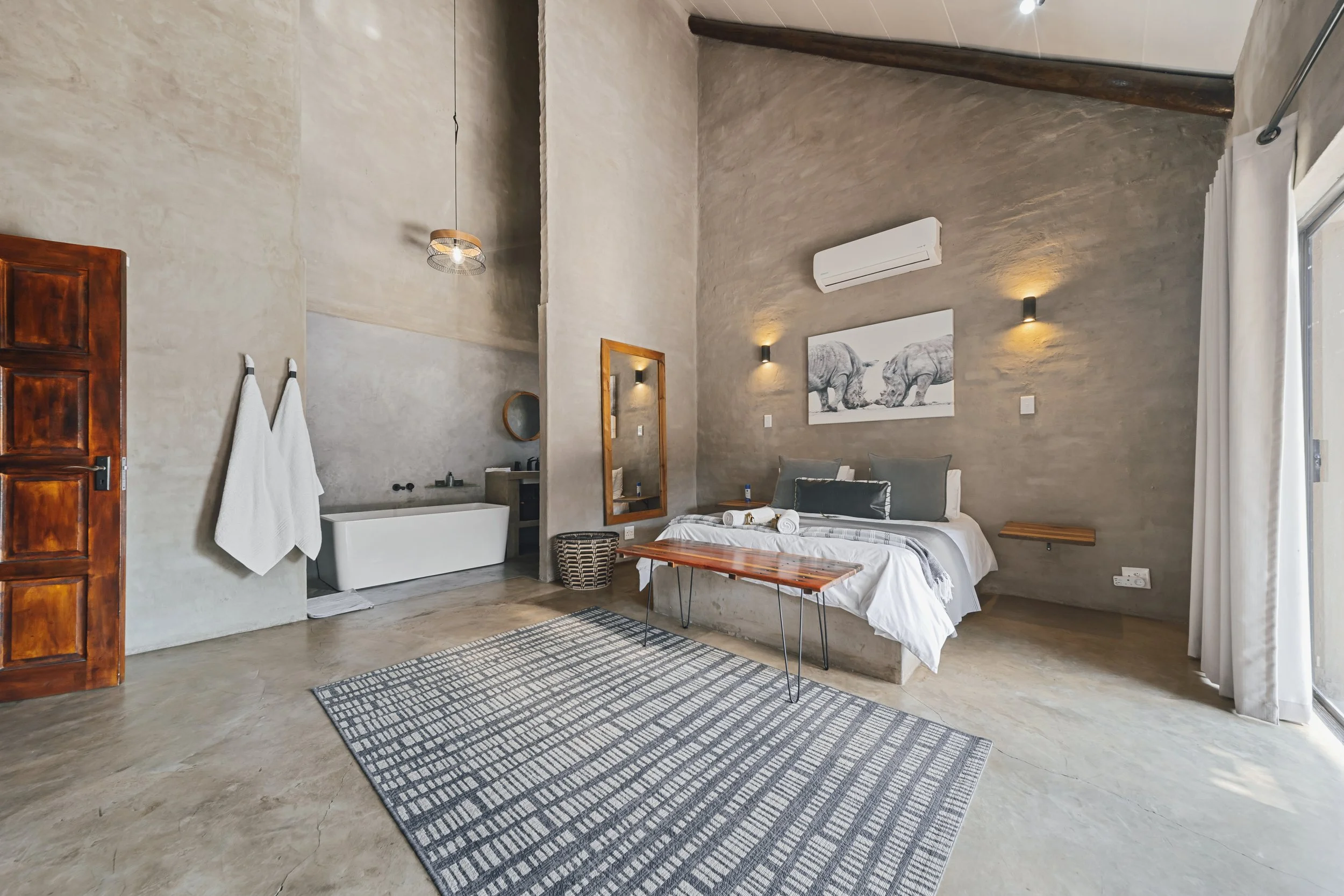 A bedroom with a high ceiling, beige textured walls, and a concrete floor. There is a double bed with white linens, gray pillows, and a wooden bench at the foot. To the left, a mirror hangs on the wall next to a small sink area. Two white towels are 