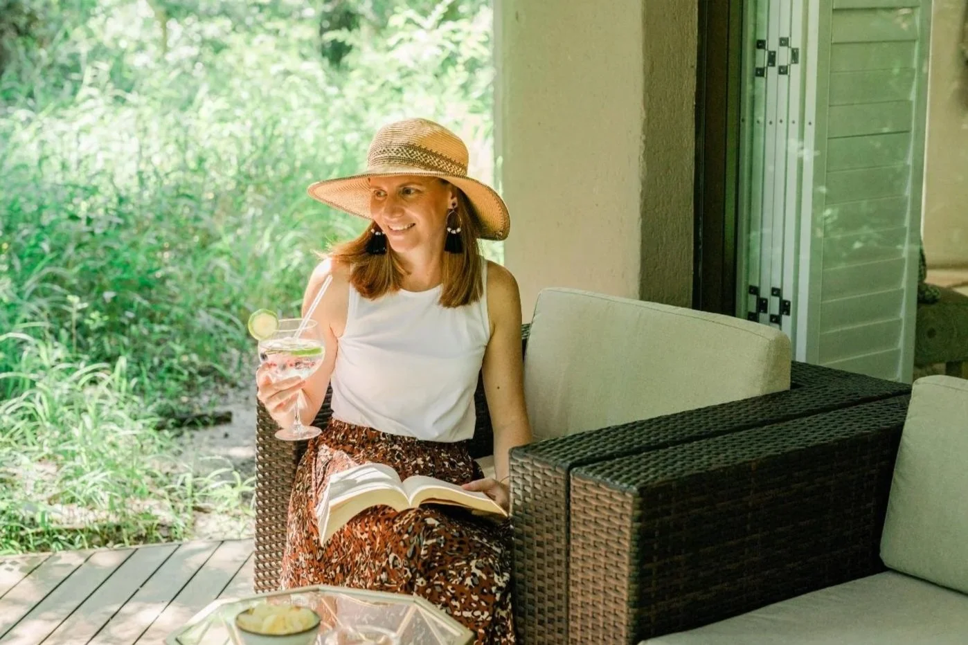 A woman sitting outdoors on a wicker chair, wearing a wide-brimmed straw hat, white sleeveless top, and patterned skirt, smiling while holding a glass of cocktail with lime and cucumber slices, and reading a book, surrounded by greenery and patio fur