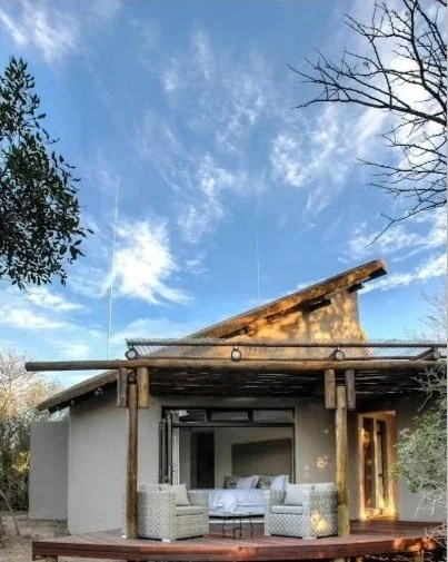 A cozy outdoor patio with a view of a modern house, featuring wicker chairs, a bed, and a wooden deck, with a bright blue sky and trees in the background.